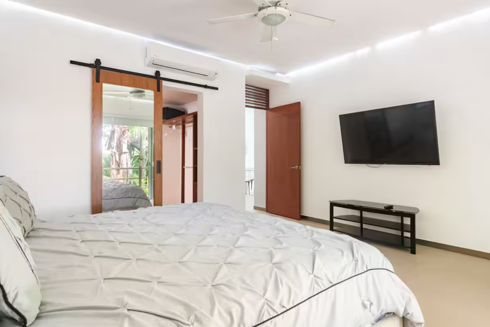 Modern bedroom with a large bed, wall-mounted TV, ceiling fan, sliding door with mirror, and lighted ceiling.