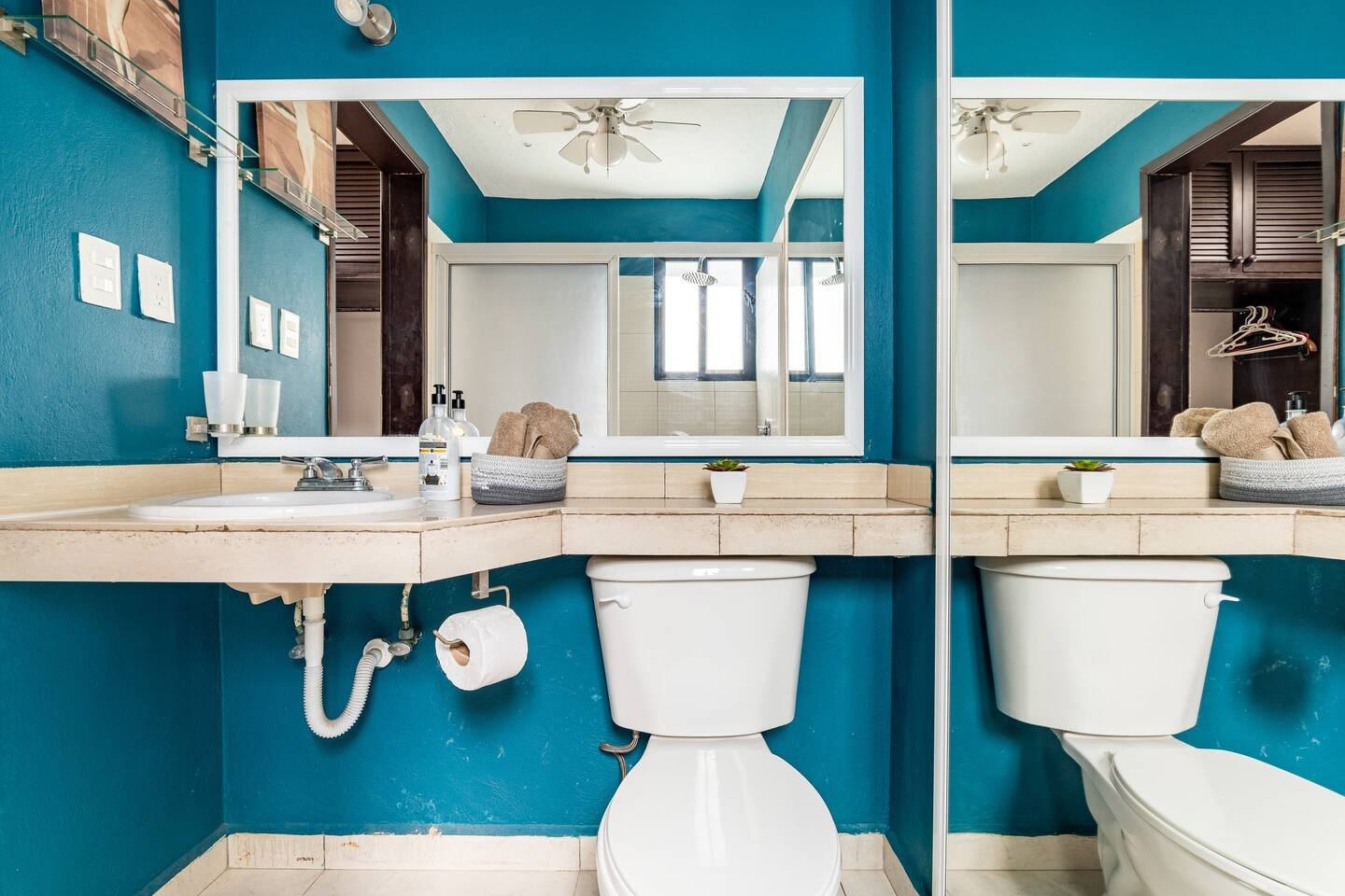 Modern bathroom with teal walls, white toilet, and a beige countertop featuring a sink, soap dispensers, and a round basket with towels. A large mirror reflects the ceiling fan, and there's a small potted plant on the counter. A toilet paper roll hol
