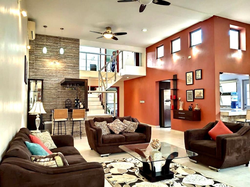 Modern living room with brown sofas, colorful cushions, glass coffee table, and a patterned rug. There's a staircase with metal railing, brown accent wall, ceiling fan, and pendant lights. Kitchen area visible in the background.