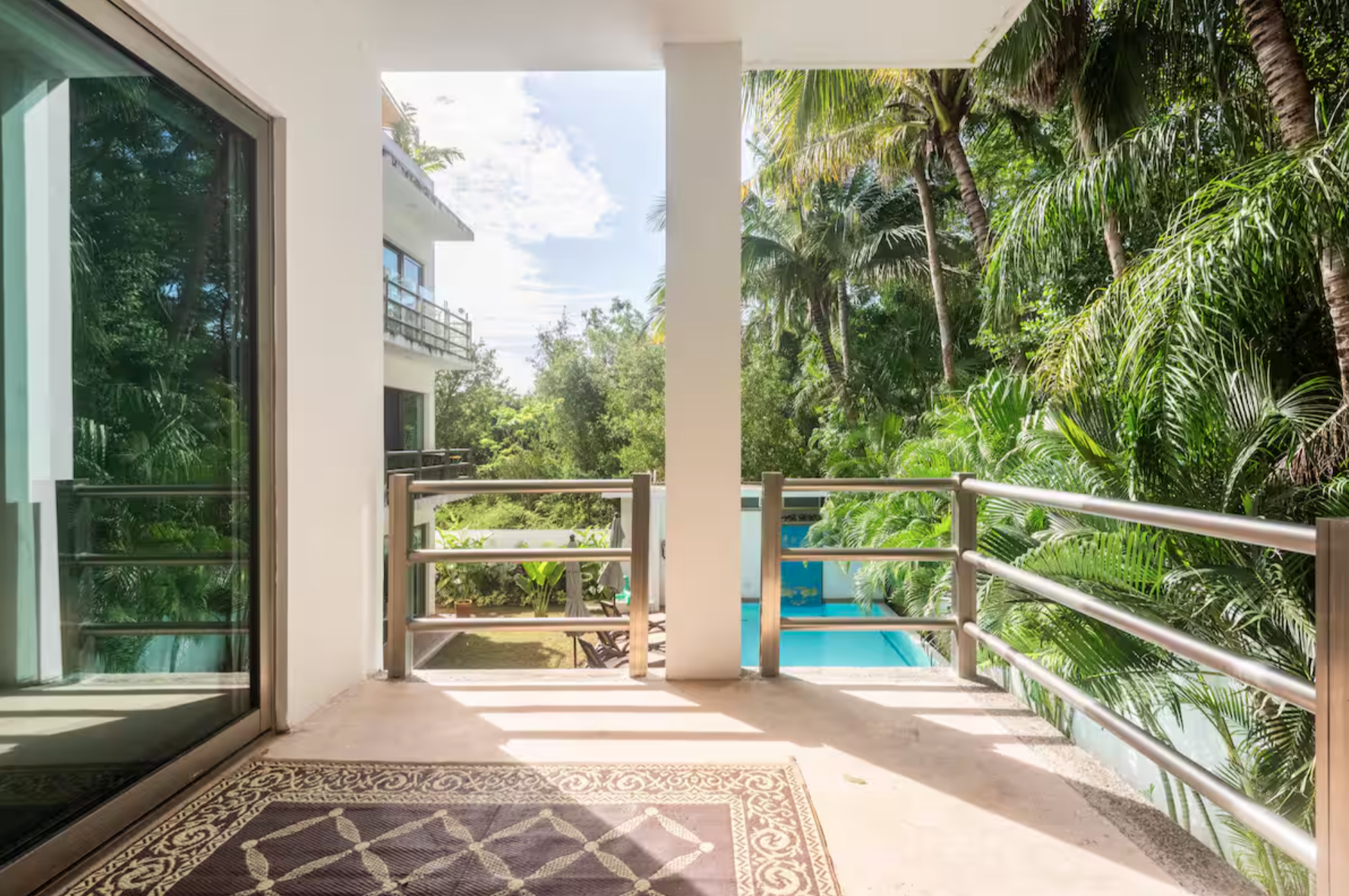 A balcony with a railing overlooks a garden with palm trees and a pool. A modern building with large windows is partially visible. The weather appears sunny with a clear sky.
