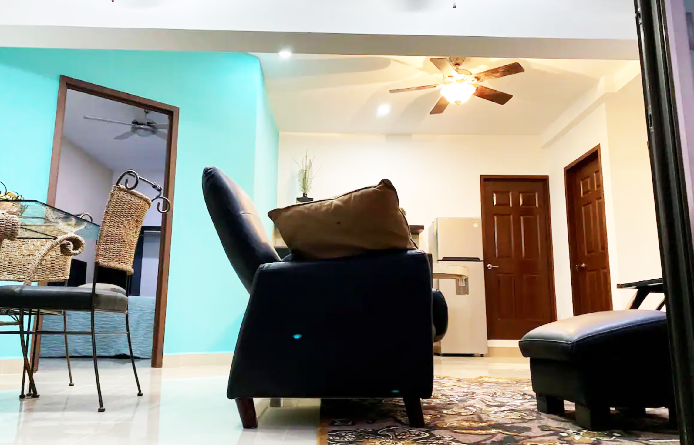 Contemporary living room with blue accent wall, black sofa, decorative pillows, ceiling fan, dining area with wicker chairs, ornate rug, and wooden doors.