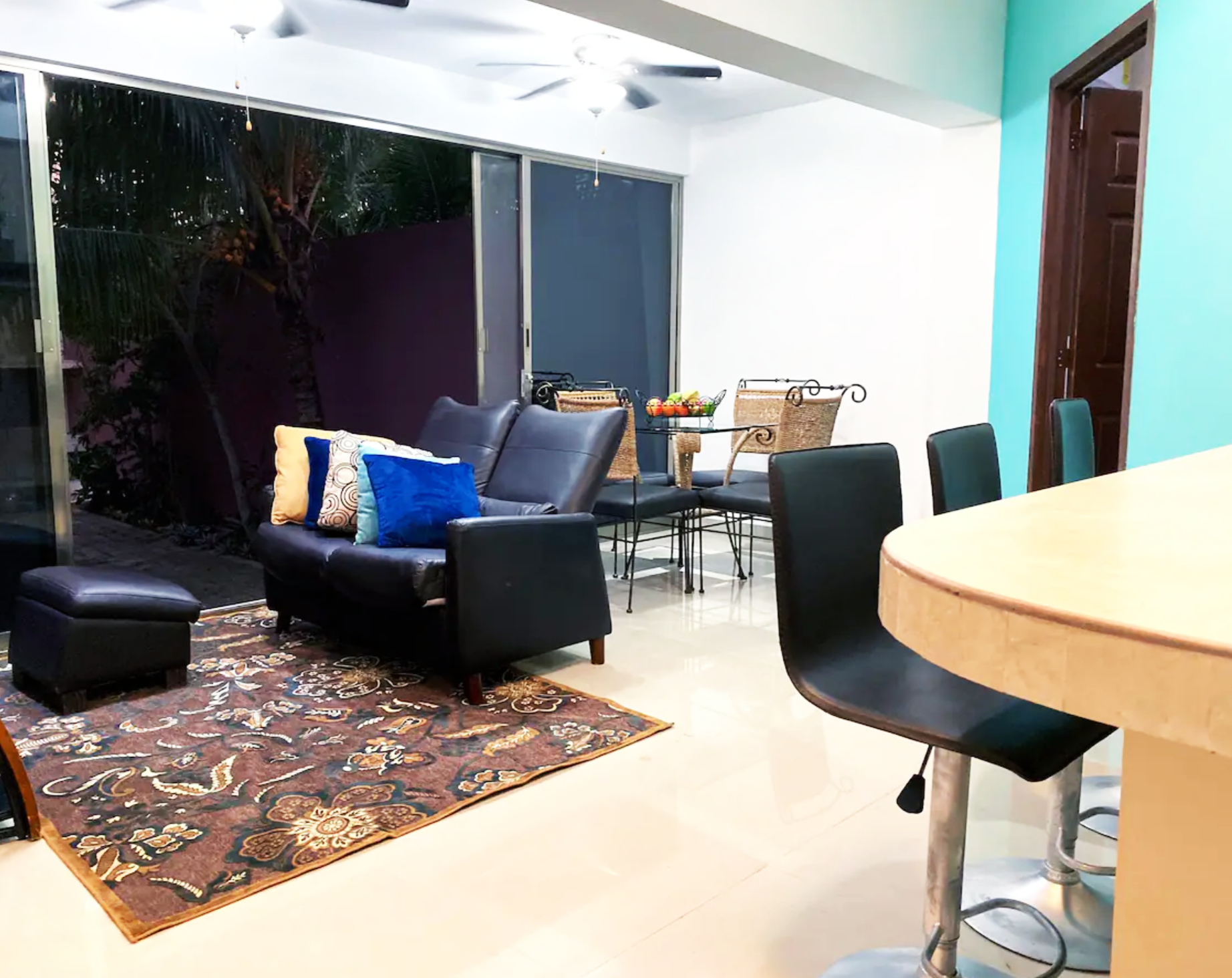 Modern living room with black leather couch, colorful pillows, decorative rug, glass dining table, and bar stools in a bright, airy setting.
