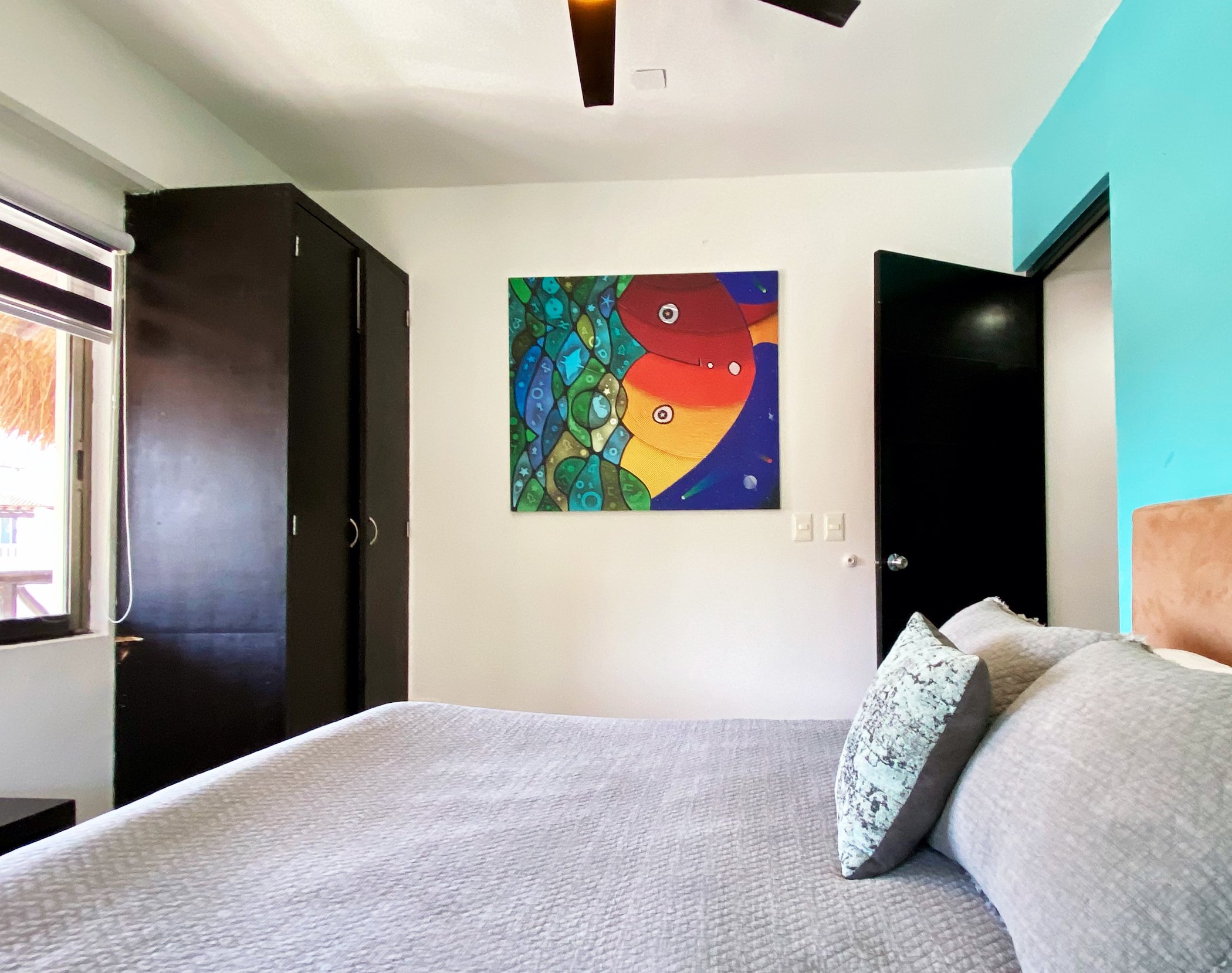 Bedroom interior with gray bedding, a colorful abstract painting on the wall, dark wardrobe, open door, ceiling fan, and natural light coming through the window.
