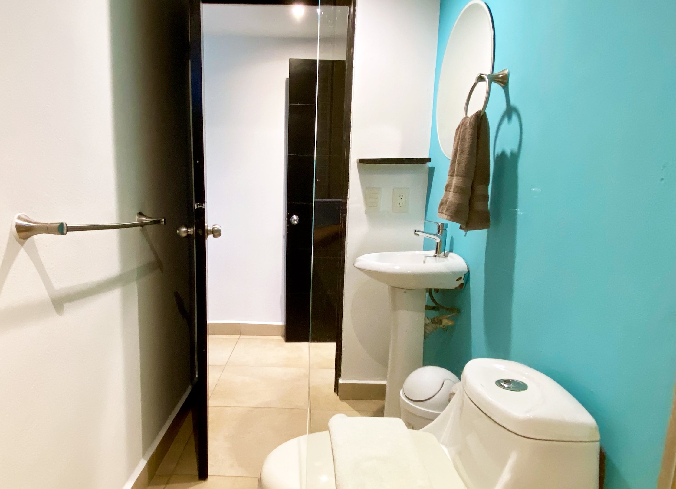 Small bathroom with turquoise wall, pedestal sink, toilet, round mirror, towel ring, and open black door.