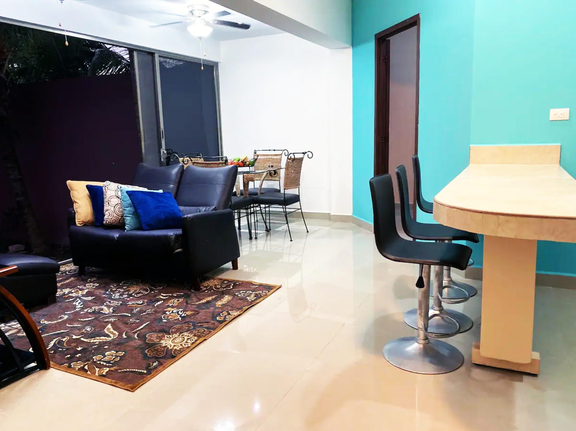 Interior of a living and dining area with a brown sofa, colorful cushions, patterned rug, modern bar stools, dining table with chairs, ceiling fan, and teal accent wall.