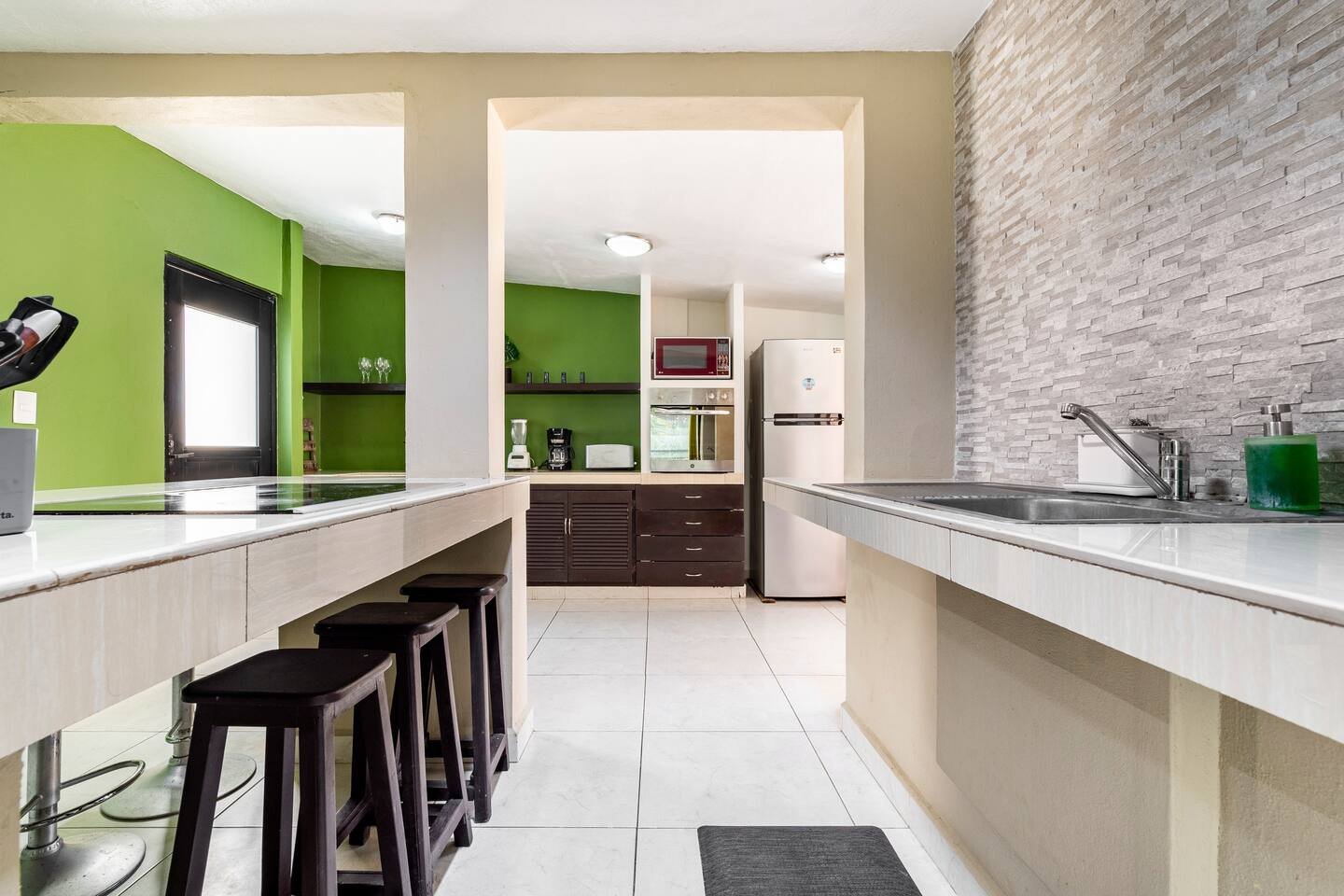 Modern kitchen with green walls, white countertops, and bar stools. Features a refrigerator, microwave, blender, coffee maker, and sink. Gray tile backsplash adds contrast.
