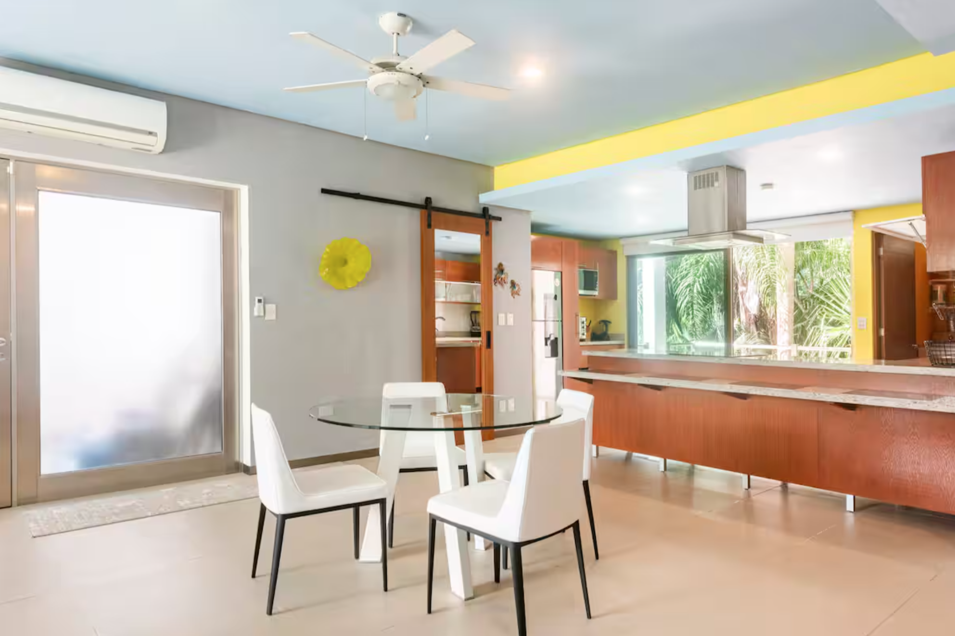 Modern kitchen and dining area with white chairs, a glass table, ceiling fan, sliding barn door, stainless steel appliances, and large window with outdoor view.