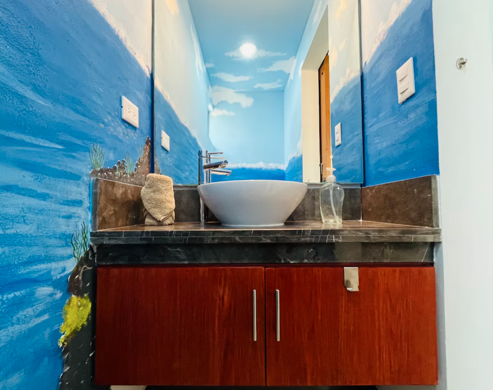 Bathroom with blue ocean-themed mural, vessel sink on marble countertop, wooden cabinet, wall-mounted faucet, and soap dispenser.