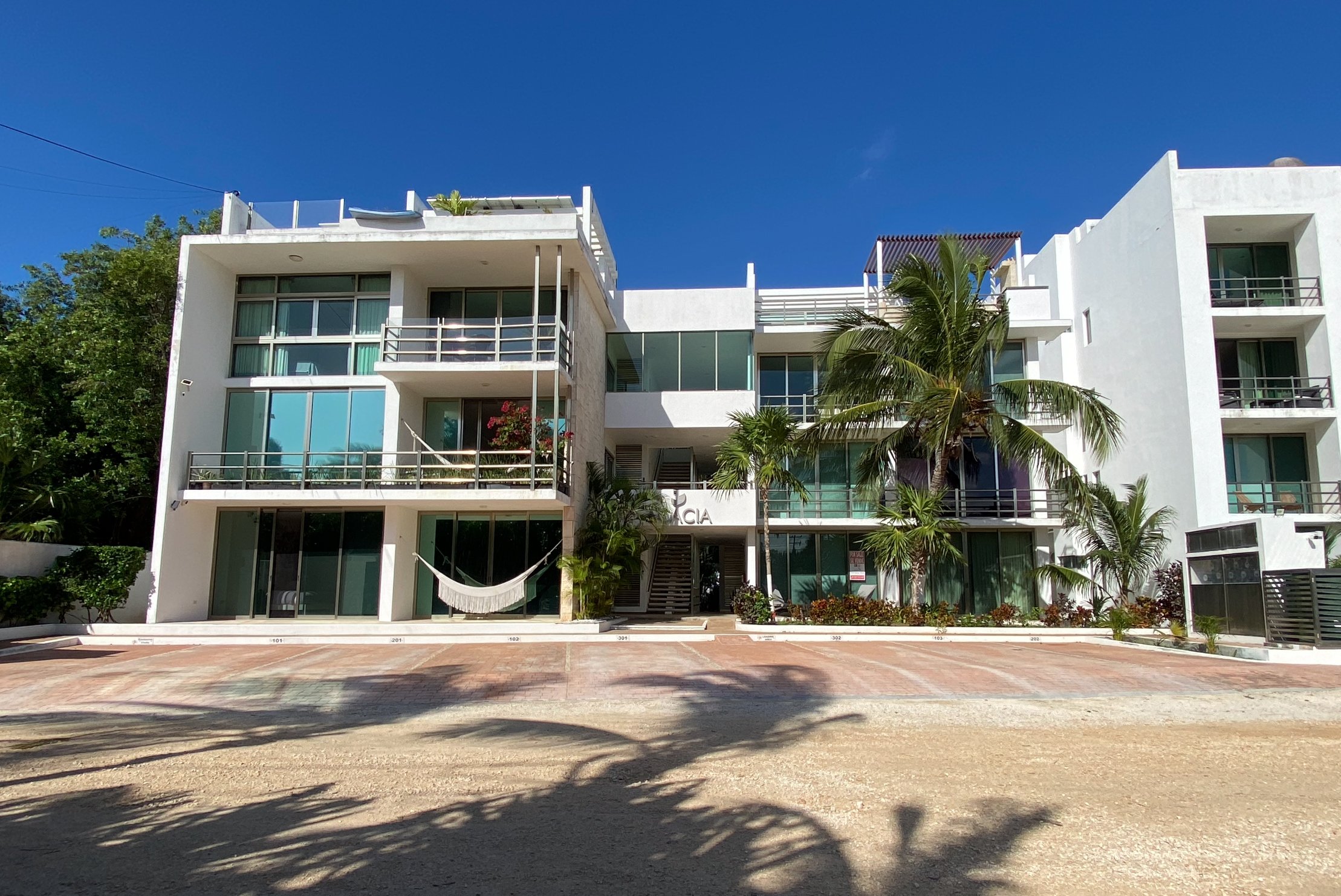 Modern white apartment building with balconies, large windows, and a hammock; surrounded by palm trees and plants under a blue sky.