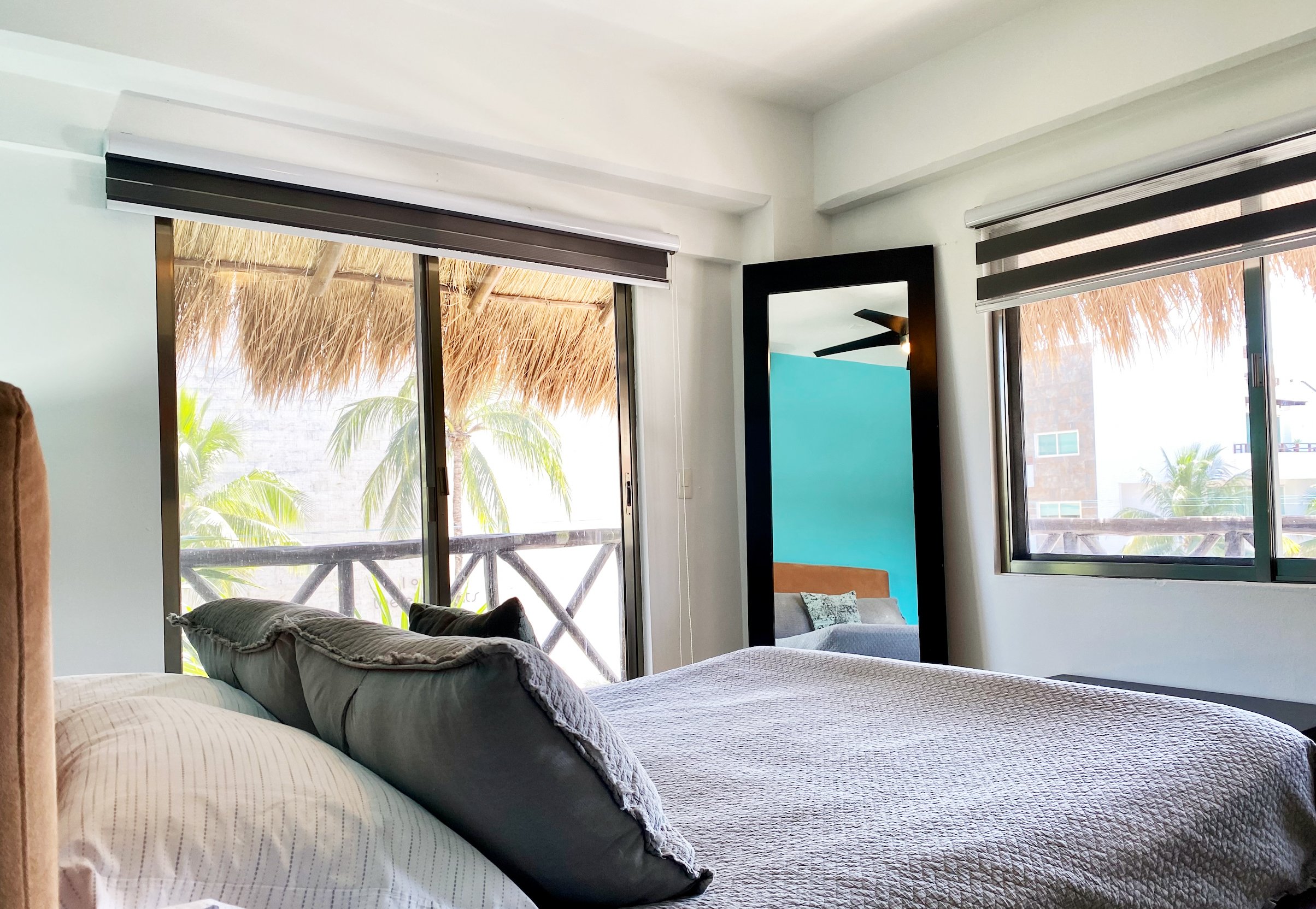 Bedroom with a bed, large cushion, window view of palm trees, and a mirror reflecting part of a turquoise wall and ceiling fan.