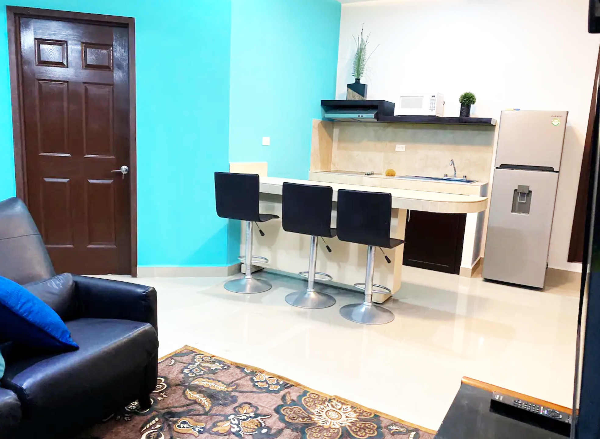Modern kitchen and living area with turquoise walls, brown door, black leather chair, patterned rug, kitchen counter with three bar stools, stainless steel refrigerator, microwave, and decorative plants.