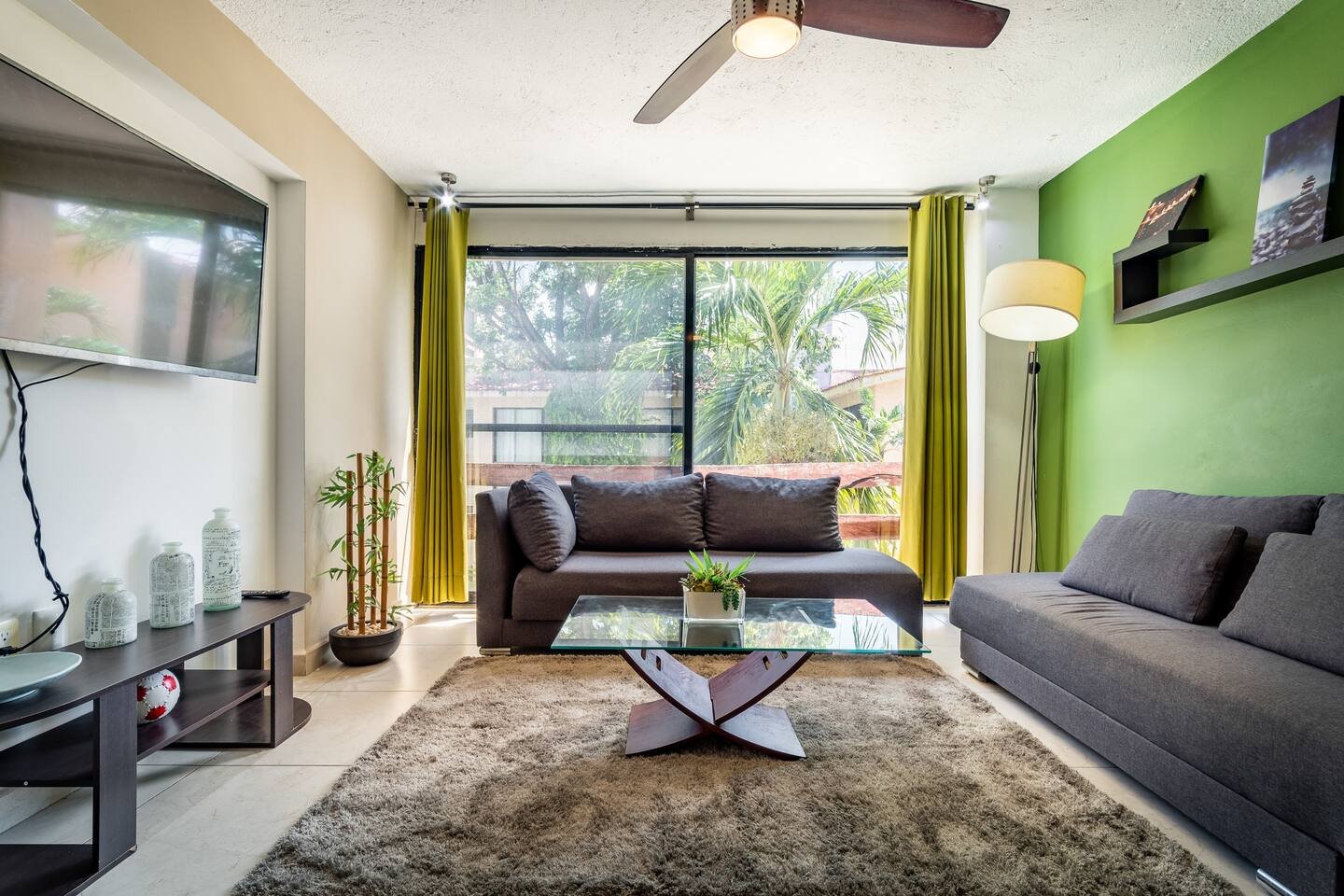 Modern living room with gray sofas, glass coffee table, green accent wall, flat-screen TV, yellow curtains, rug, and floor lamp. A large window showcases outdoor greenery.