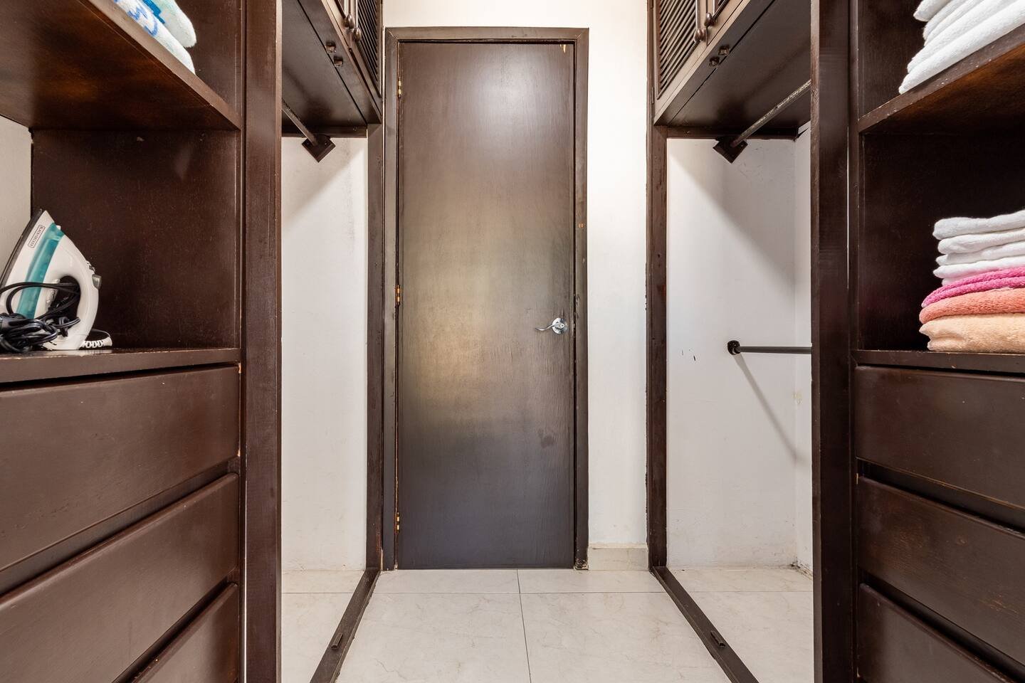 Walk-in closet with dark wood shelves containing folded towels and an iron, and a central closed door at the back.