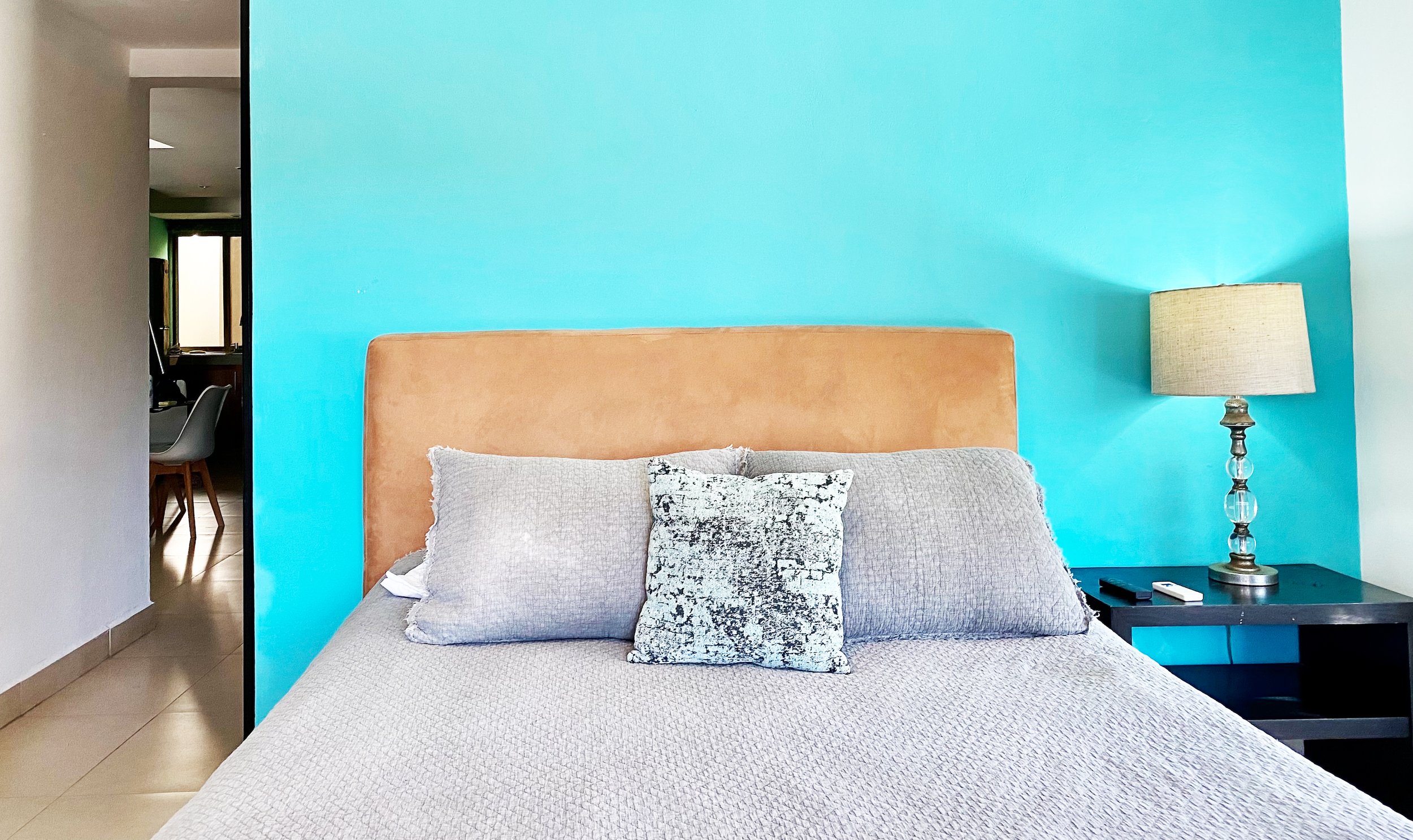 Bedroom with a bed featuring gray bedding and pillows against a turquoise wall, a bedside lamp on a nightstand, and an open entryway to another room.
