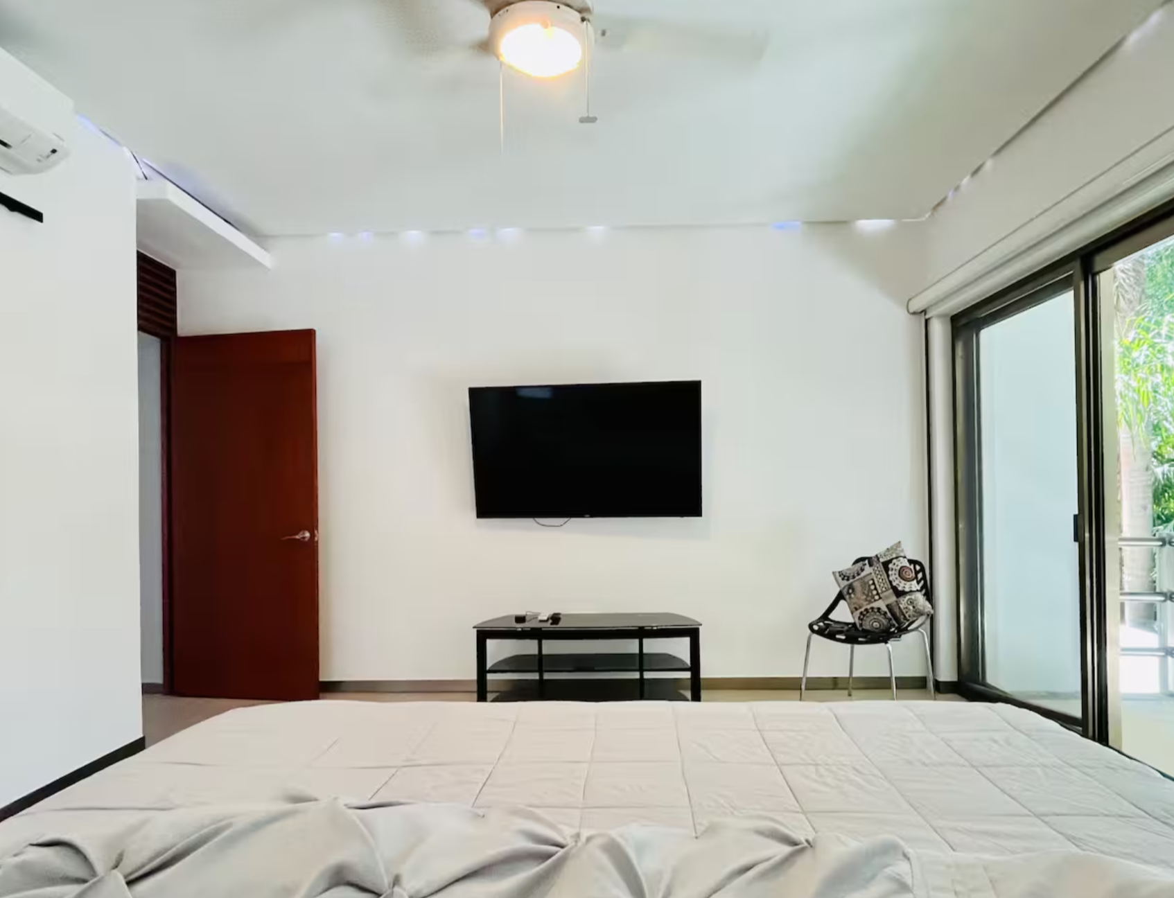Minimalist bedroom with a ceiling fan, wall-mounted TV, wooden door, and sliding glass doors leading to a balcony.