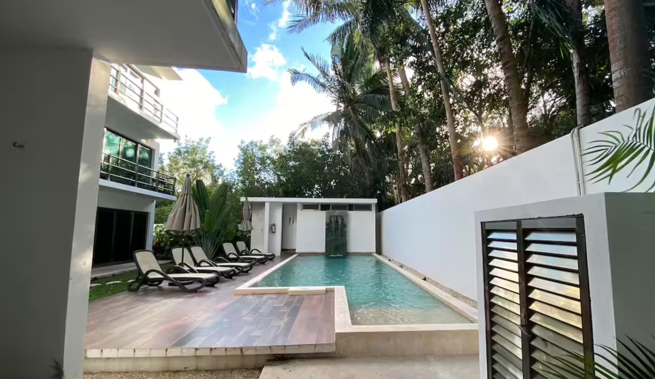 Modern outdoor swimming pool area with lounge chairs and umbrellas, surrounded by tall trees and a blue sky background.