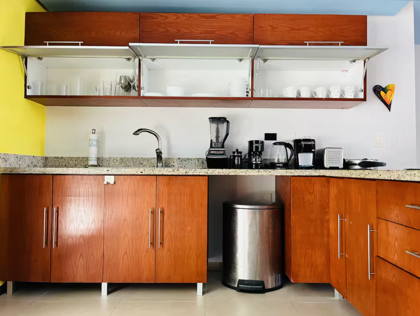 Modern kitchen with wooden cabinets, open shelving, countertop appliances, and a stainless steel trash can.