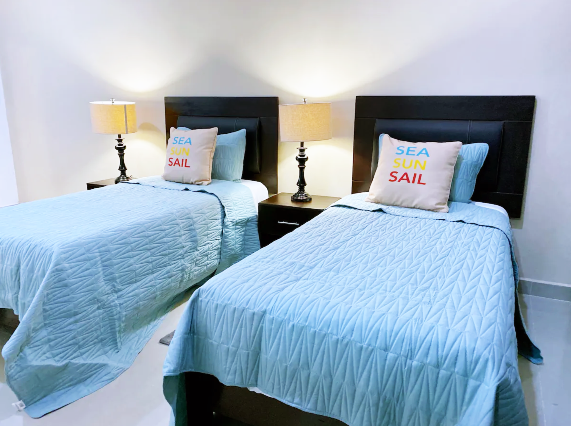 Twin beds with light blue quilts, decorative pillows with 'Sea Sun Sail' text, bedside tables with lamps.