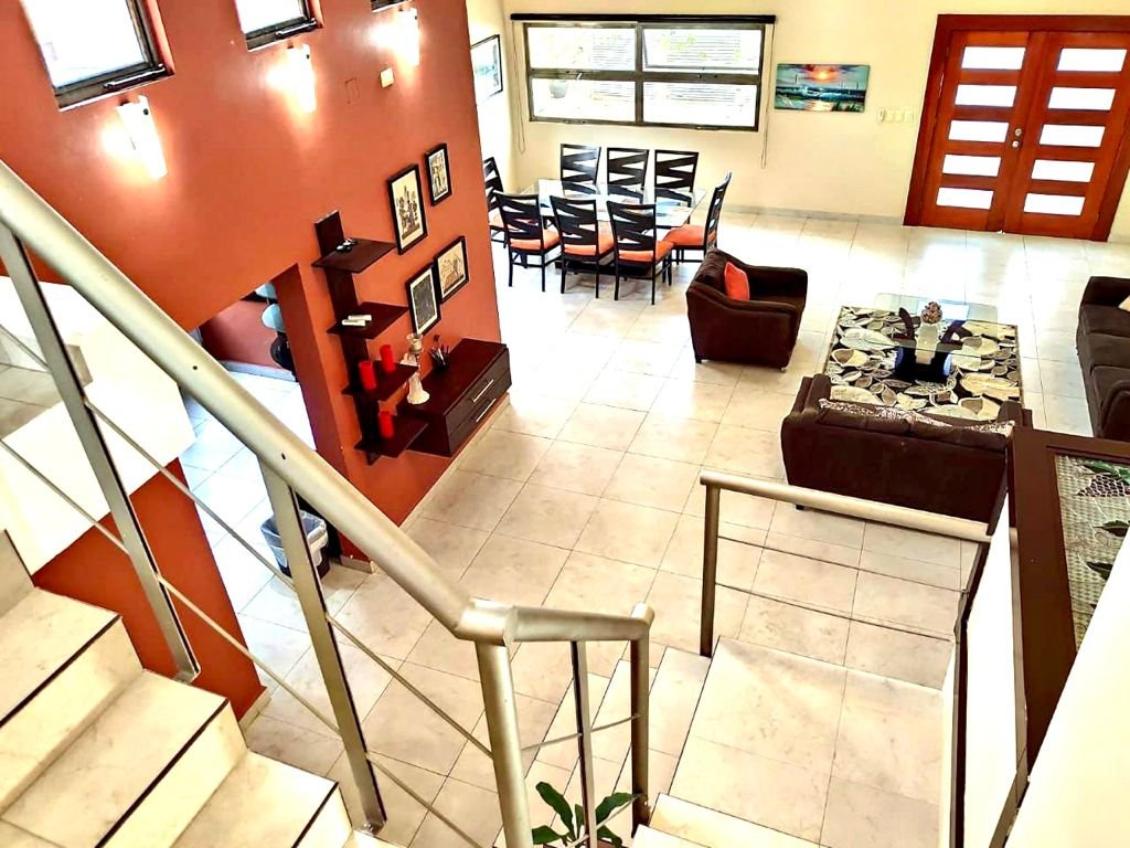 Interior of a modern living room with a staircase, a dining table, and sofas, featuring a red accent wall, framed artwork, and decorative items.
