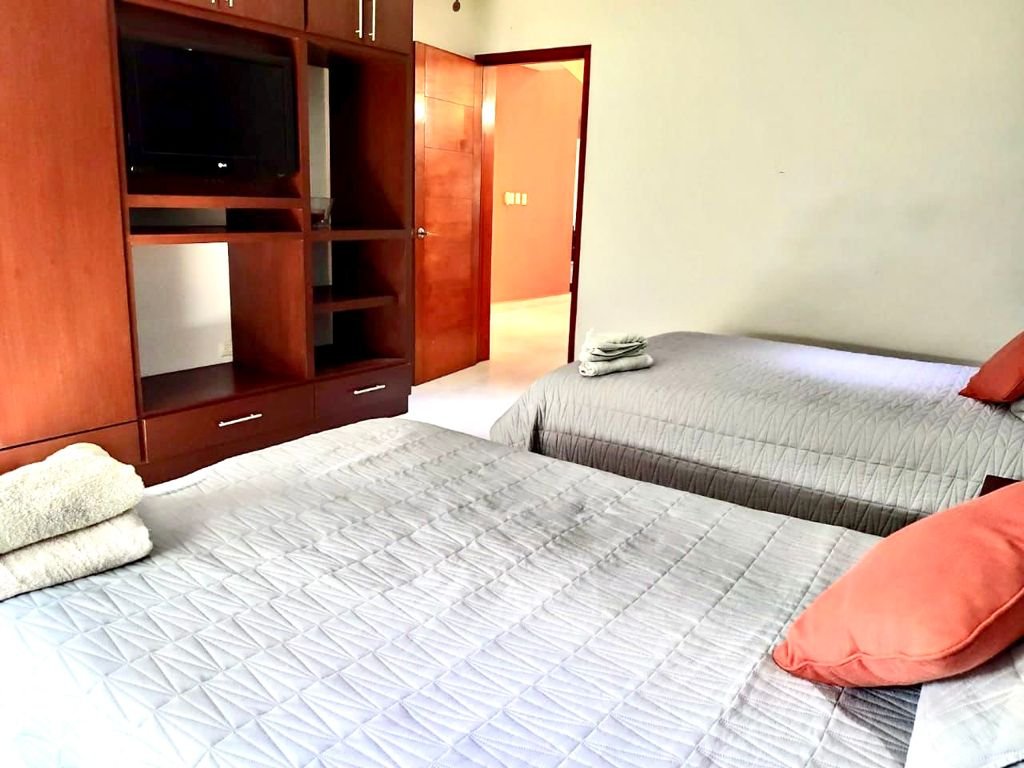 Bedroom with two neatly made beds, gray quilts, and orange accent pillows. A wooden entertainment unit with a flat-screen TV is visible. Towels are folded on the beds. A door leads to an adjacent room.