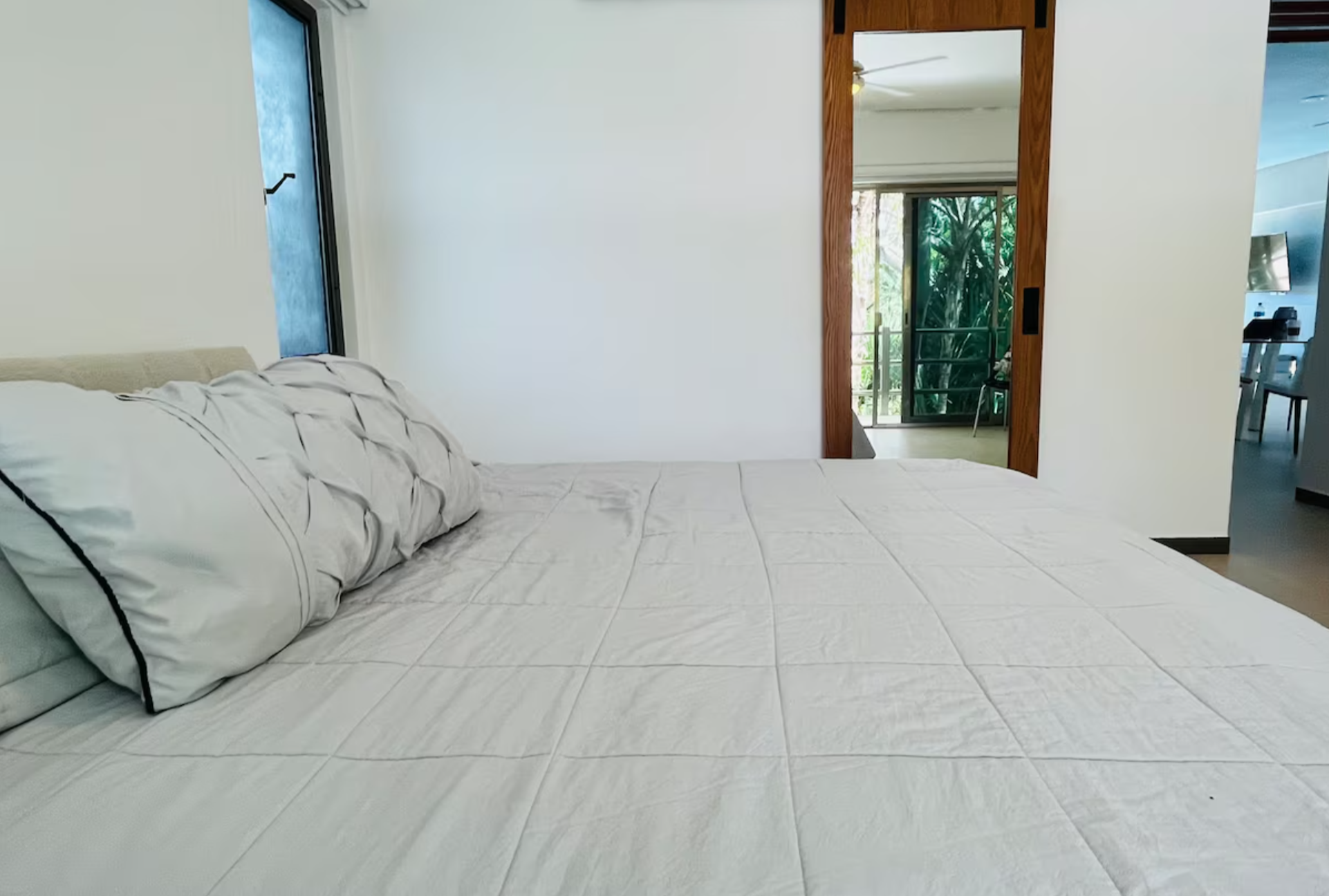 Bedroom with a neatly made bed, large pillows, white bedding, sliding wooden door, and glimpse of a balcony with greenery visible.
