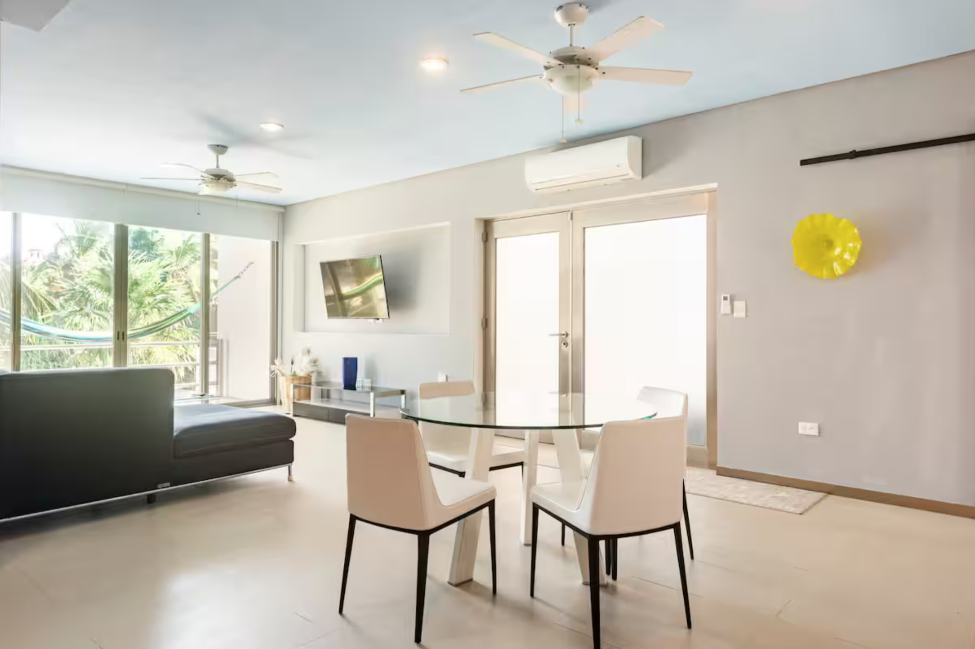 Modern living room with a glass dining table, four chairs, wall-mounted TV, large sectional sofa, ceiling fans, sliding glass doors, and a yellow decorative wall piece.