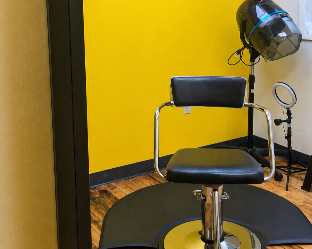 A black hair salon styling chair in front of a yellow wall, with hair salon equipment including a hooded hair dryer and ring light visible in the background.