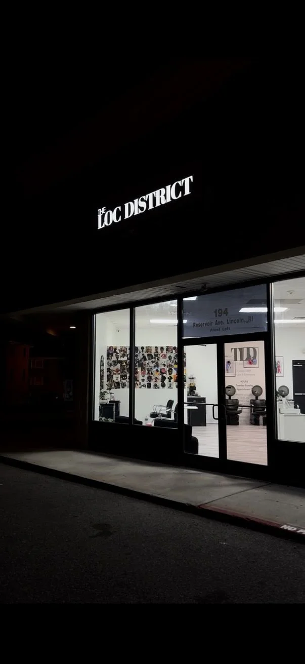 Exterior view of an office or studio with a sign that reads 'The LOC District,' showing a brightly lit interior through large glass windows, with a wall decorated with photos and some furniture.