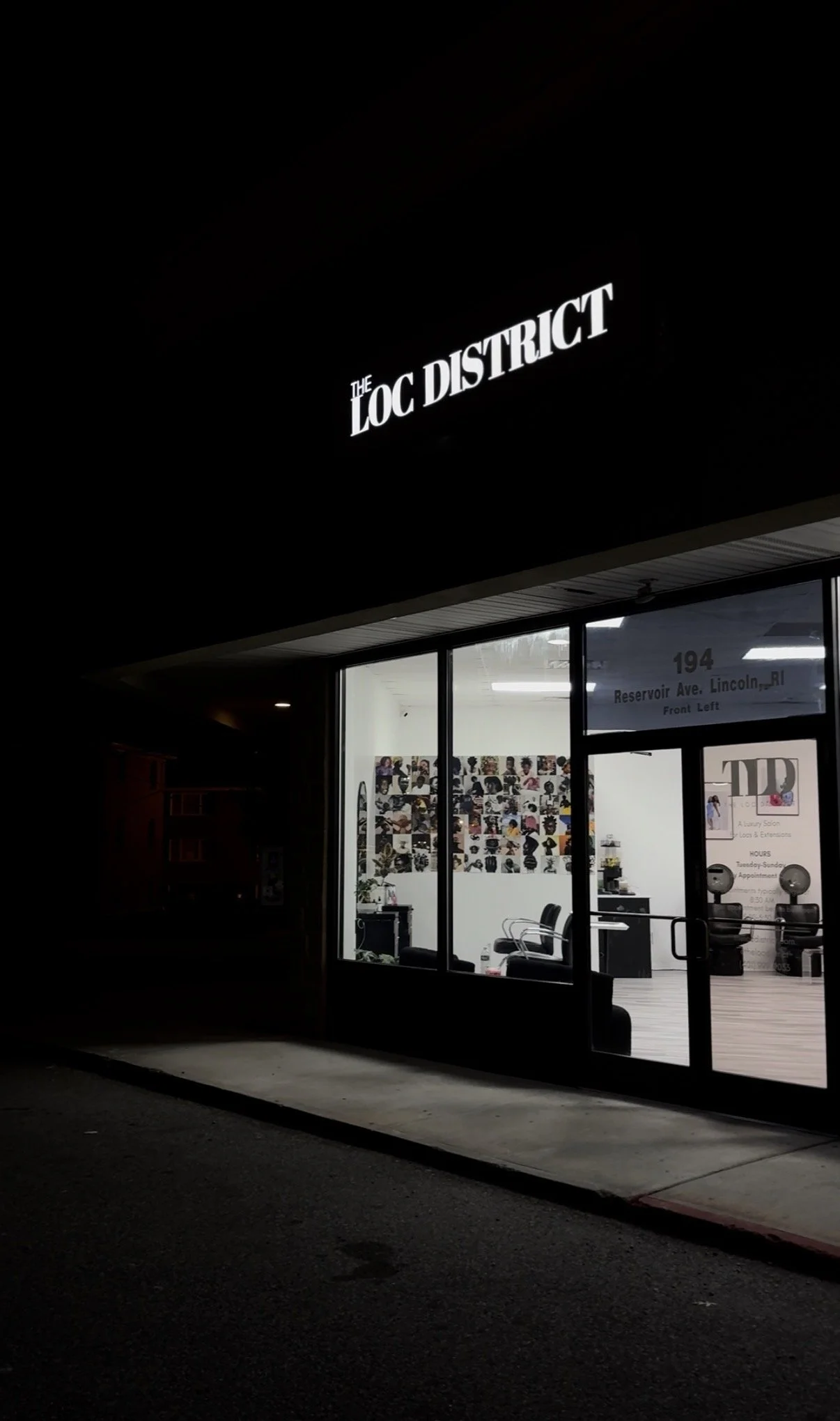 Nighttime exterior view of a hair salon named 'The LOC District' with large glass windows showcasing hair care chairs and hair styling images inside. The sign is illuminated in white on the black facade.