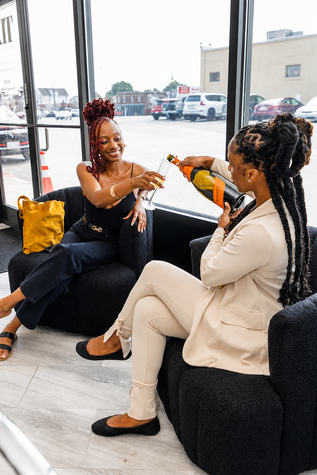 Two women sitting on black couches inside a building by a large window. One woman with red dreadlocks is toasting with a glass of champagne held by a woman with dark curly hair, dressed in a white blazer and pants. Outside the window, parked cars and a building are visible.