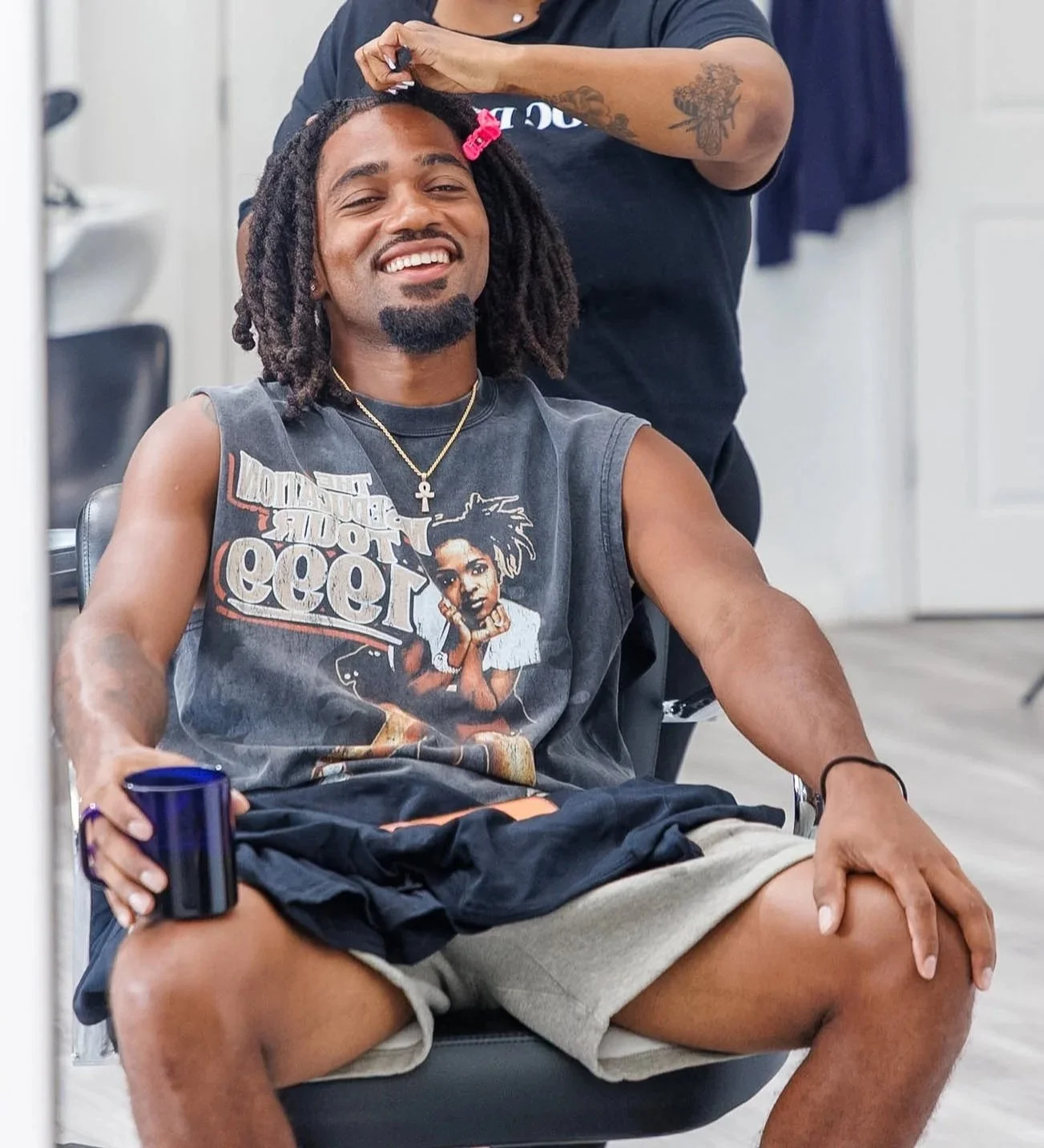 A man with dreadlocks and a big smile sitting in a salon chair, getting his hair styled by a hairdresser. The man is wearing a sleeveless graphic T-shirt and beige shorts, holding a blue cup.