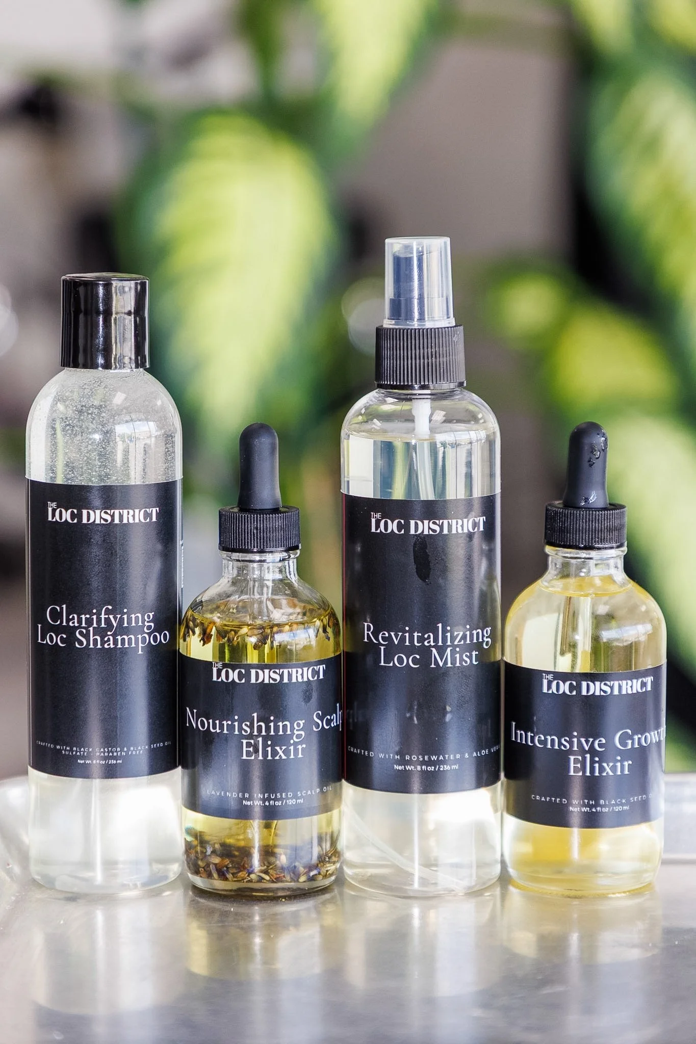 A collection of hair care products with black labels from The Loc District, including clarifying loc shampoo, nourishing scalp elixir, revitalizing loc mist, and intensive growth elixir, set against a background of blurred green leaves.