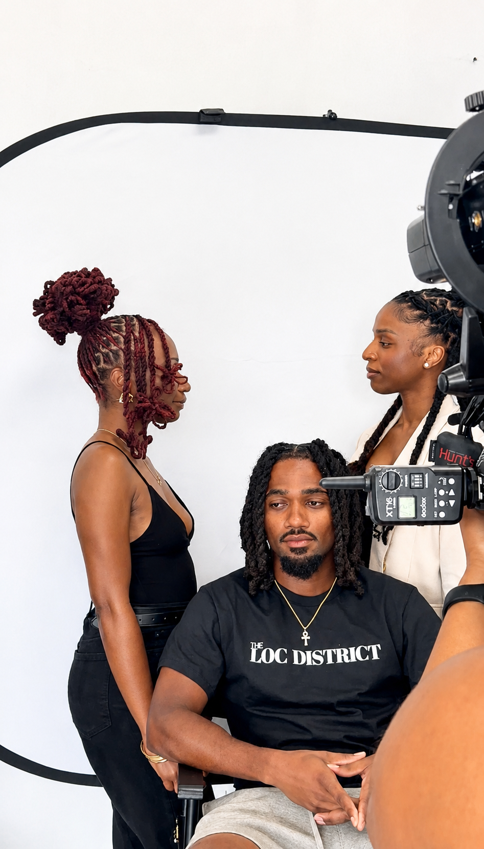 A film scene with three Black individuals, two women and one man, involved in a shoot. The man sits in front, wearing a black t-shirt that reads 'THE LOC DISTRICT' and a gold chain with an ankh pendant. One woman with styled dreadlocks is standing be
