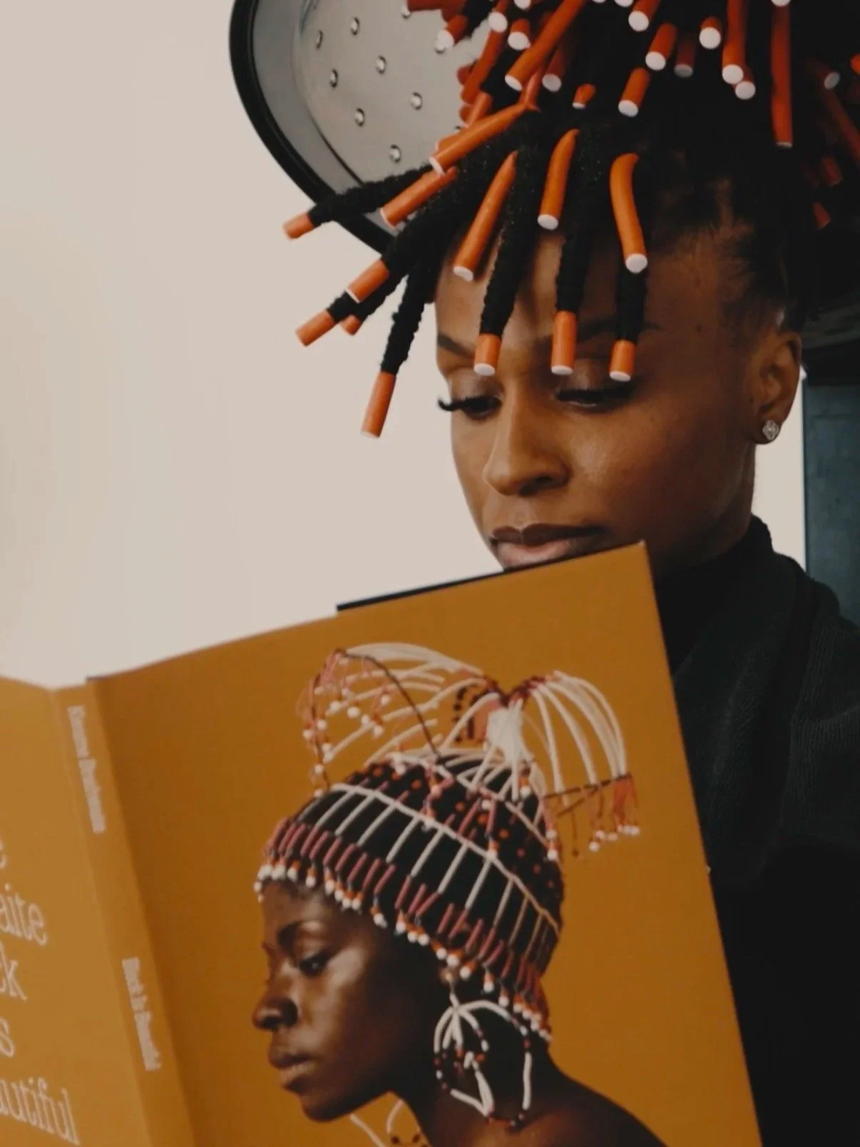 A woman with styled orange, black, and white curled hair looks at a magazine featuring a woman with similar hair styling and traditional beadwork headwear.