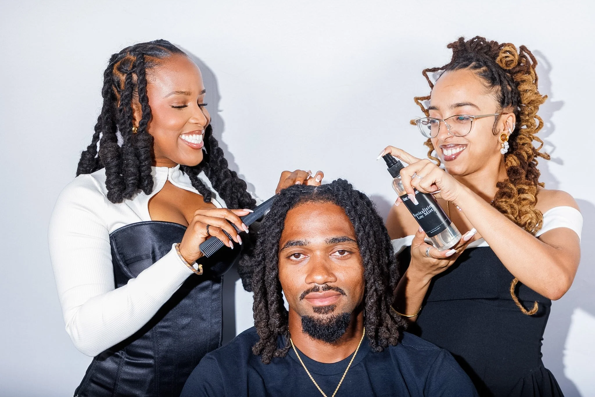Three people are grooming a man with dreadlocks. Two women, one with curly brown hair and glasses, the other with long black hair, are working on his hair with hair tools. The man has a beard and looks directly at the camera.