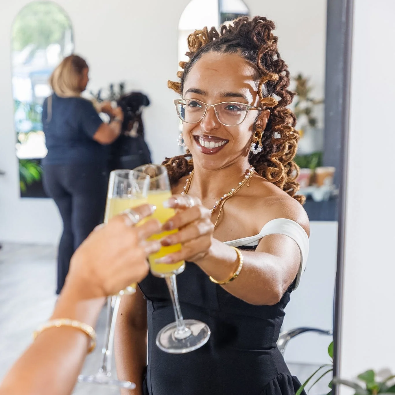 A woman with glasses and jewelry smiling as she toasts with another person holding a glass of yellow beverage, inside a bright room with a mirror behind her.