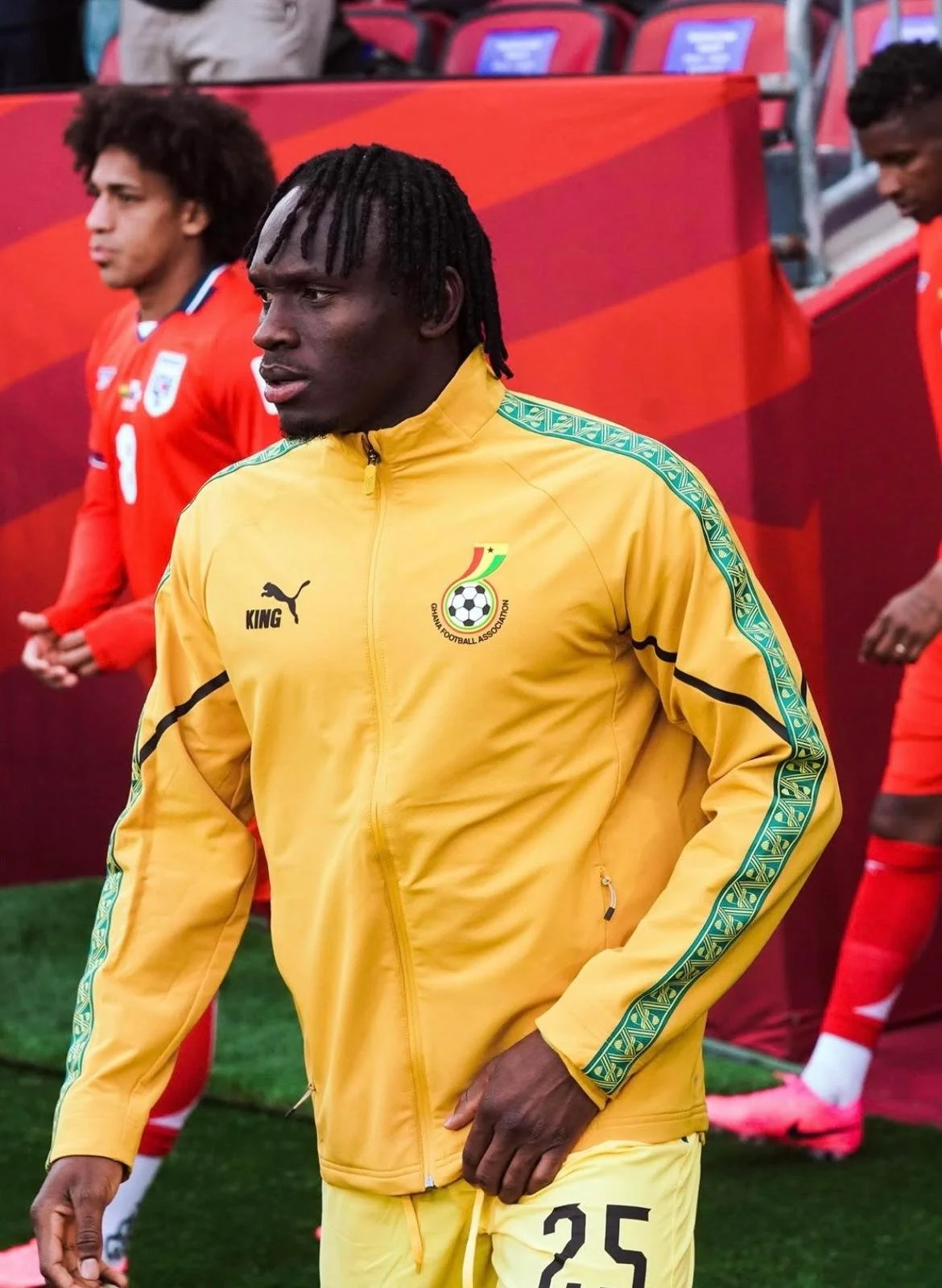 A soccer player wearing a yellow jacket with the Cameroon Football Association logo, standing on a field with other players in red jerseys in the background.