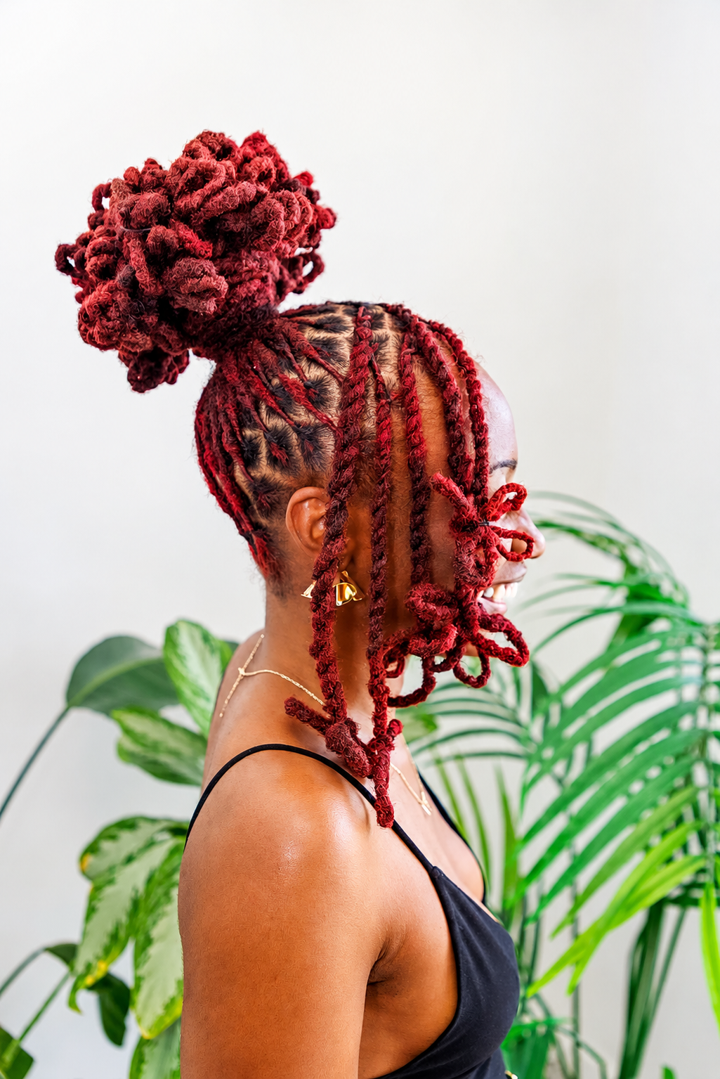 A woman with vibrant red dreadlocks styled in an updo hairstyle and intricate braids, wearing a black spaghetti strap top and gold jewelry, standing in front of green plants.