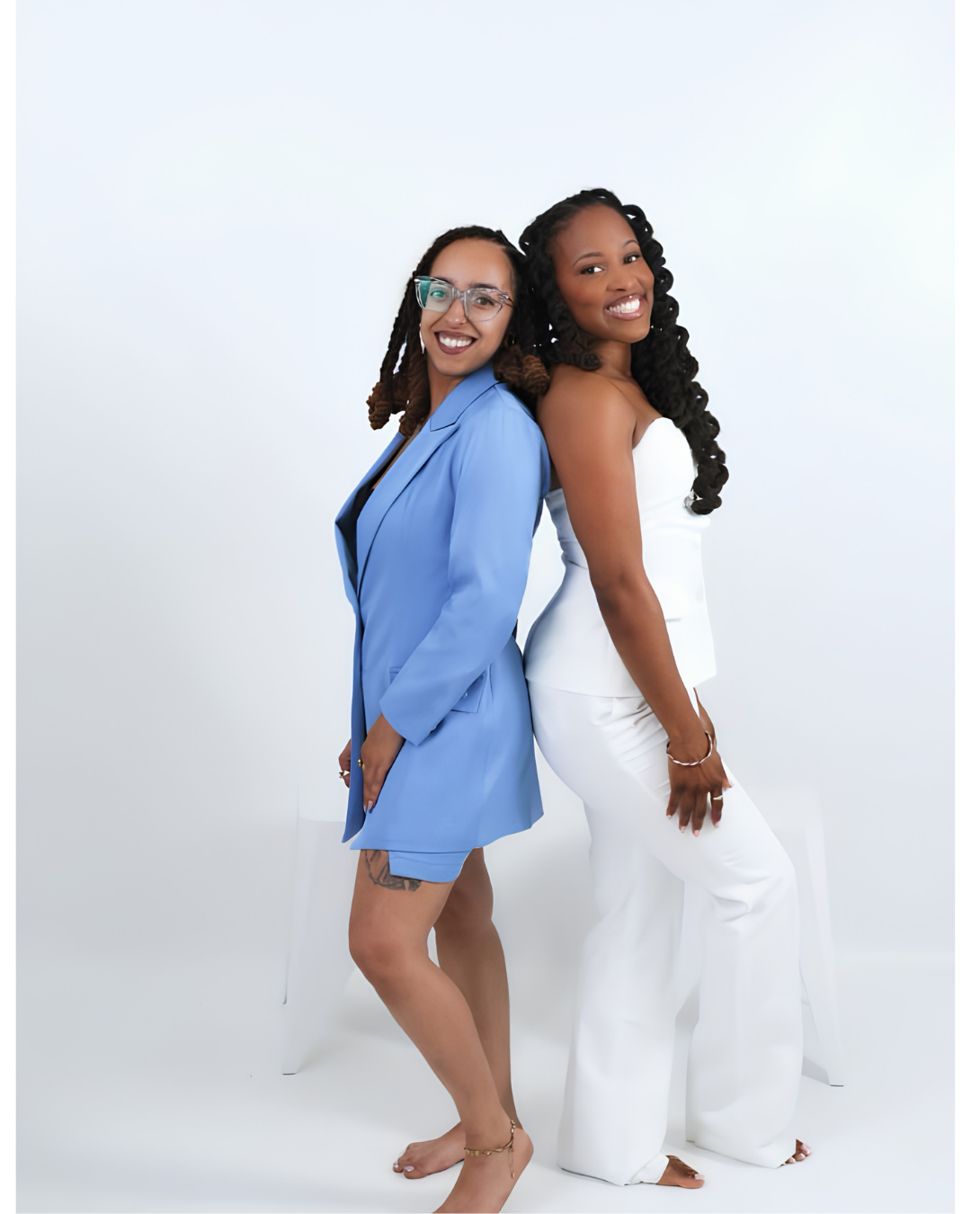 Two women standing back-to-back, smiling, with white background. One wears a blue blazer and shorts, glasses, and has dreadlocks. The other wears a white strapless jumpsuit, has long curly hair, and is barefoot.