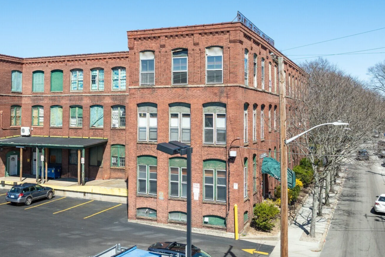A multi-story red brick building with many windows and a parking lot in front, featuring street lamps, leafless trees, and a clear blue sky.