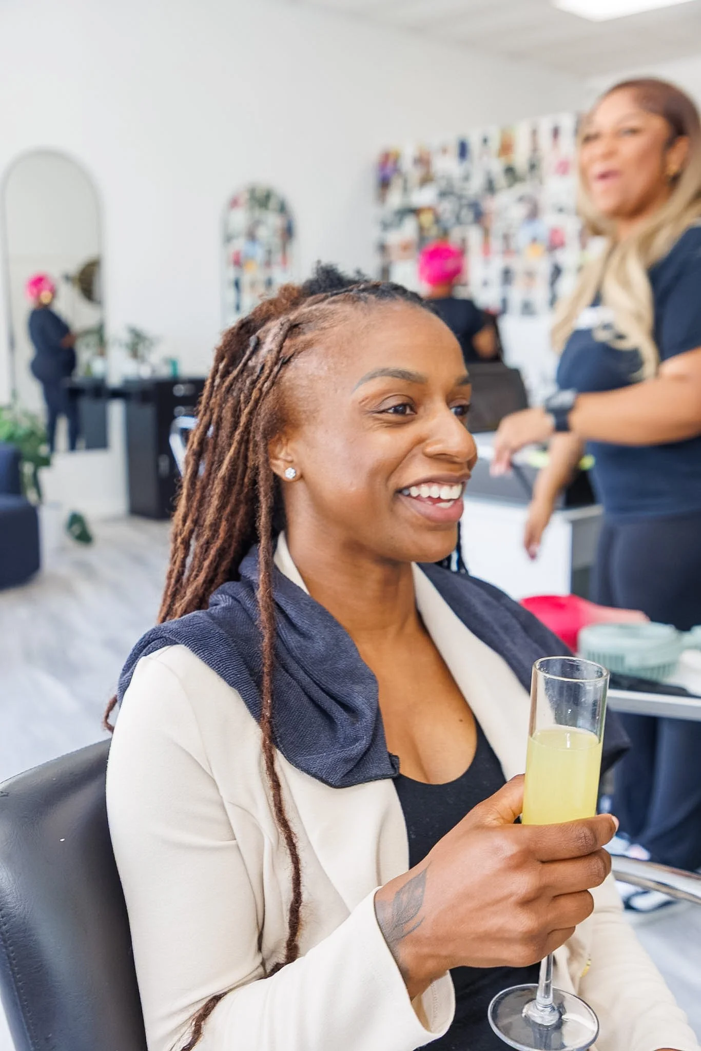 A woman with long dreadlocks and earrings smiling while holding a glass of yellow beverage in a social setting with other people and photo wall in the background.