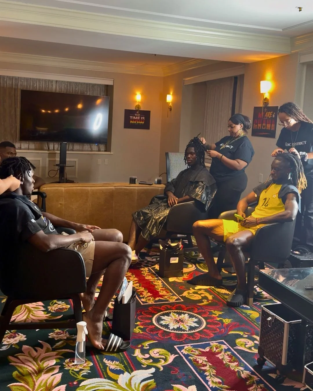 A group of young men with dreadlocks sitting in chairs in a hotel room, getting their hair styled by women standing behind them. The room has a colorful patterned carpet, a large television on the wall, and framed art and lights on the walls.