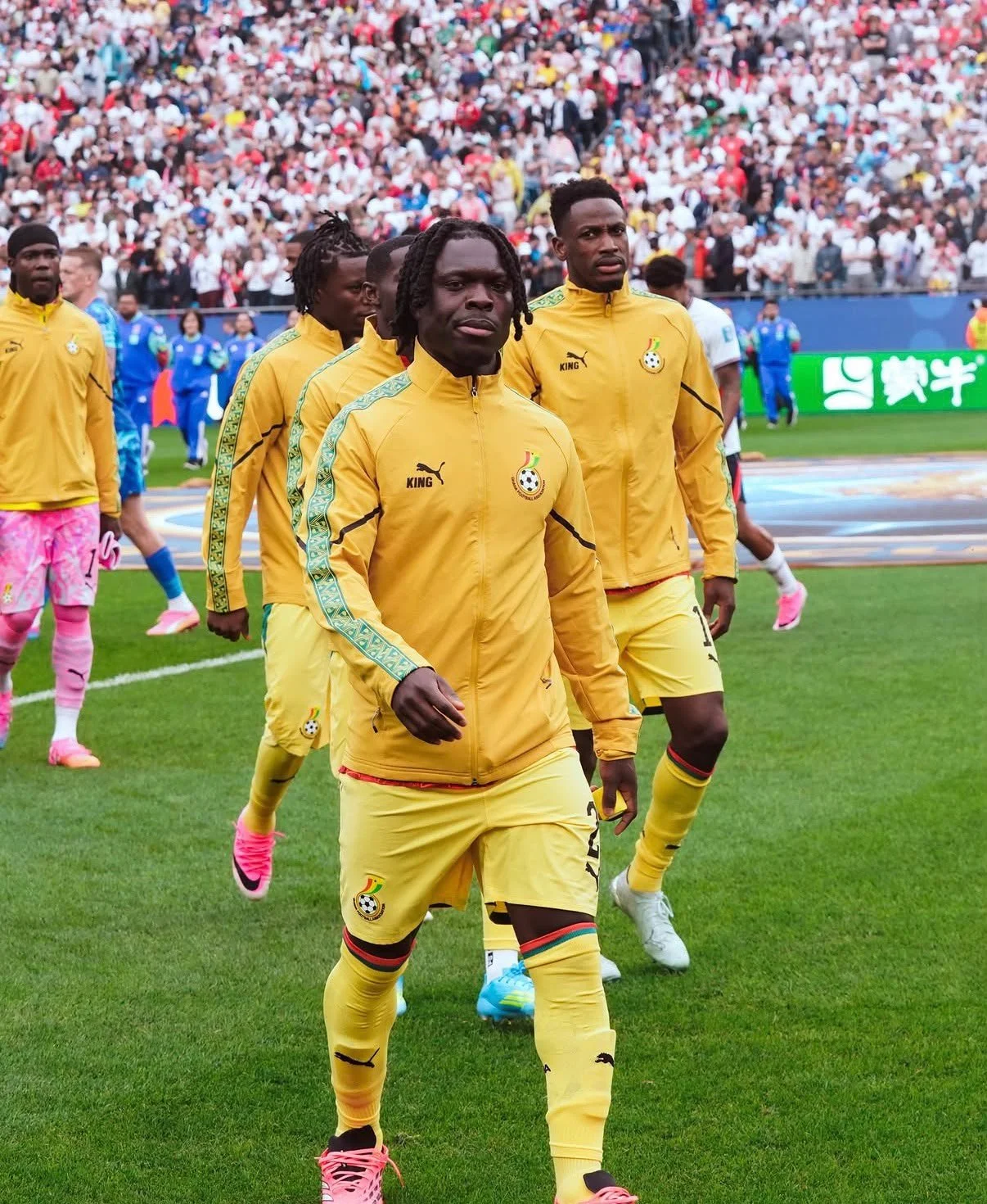 African soccer players in yellow uniforms walking on the field before a match, with a crowded stadium in the background.