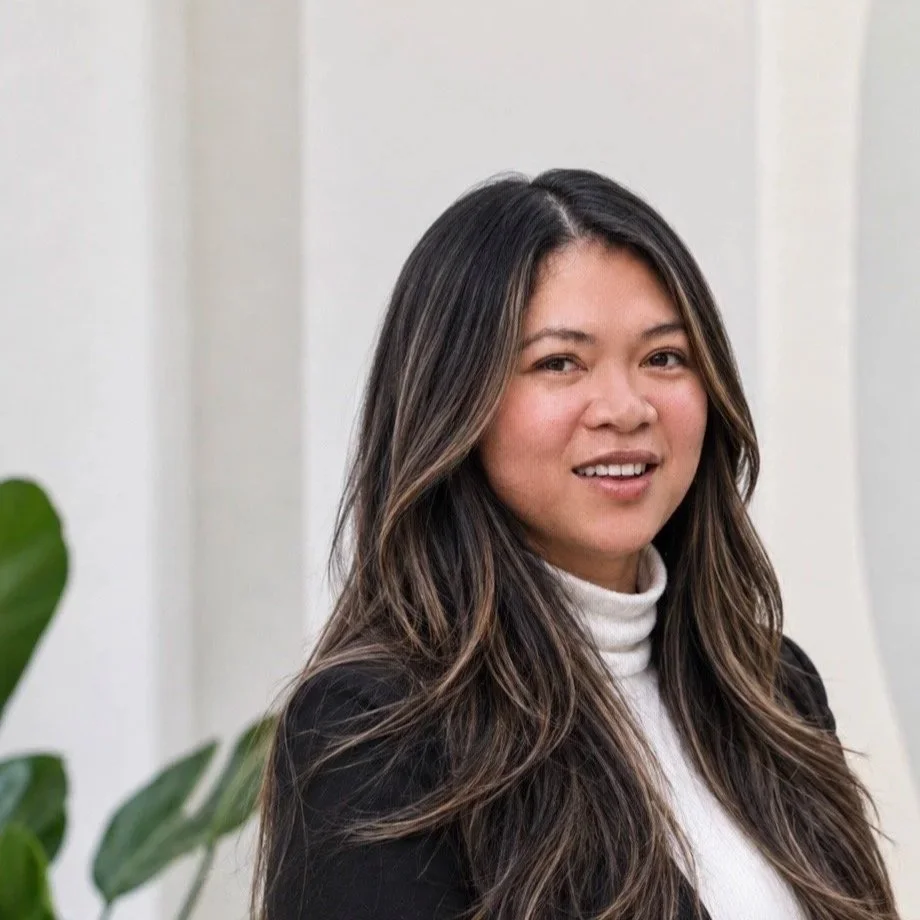 portrait of a woman with long dark hair and highlights, wearing a white turtleneck and a black blazer, standing indoors with a white wall and green plant leaves in the background