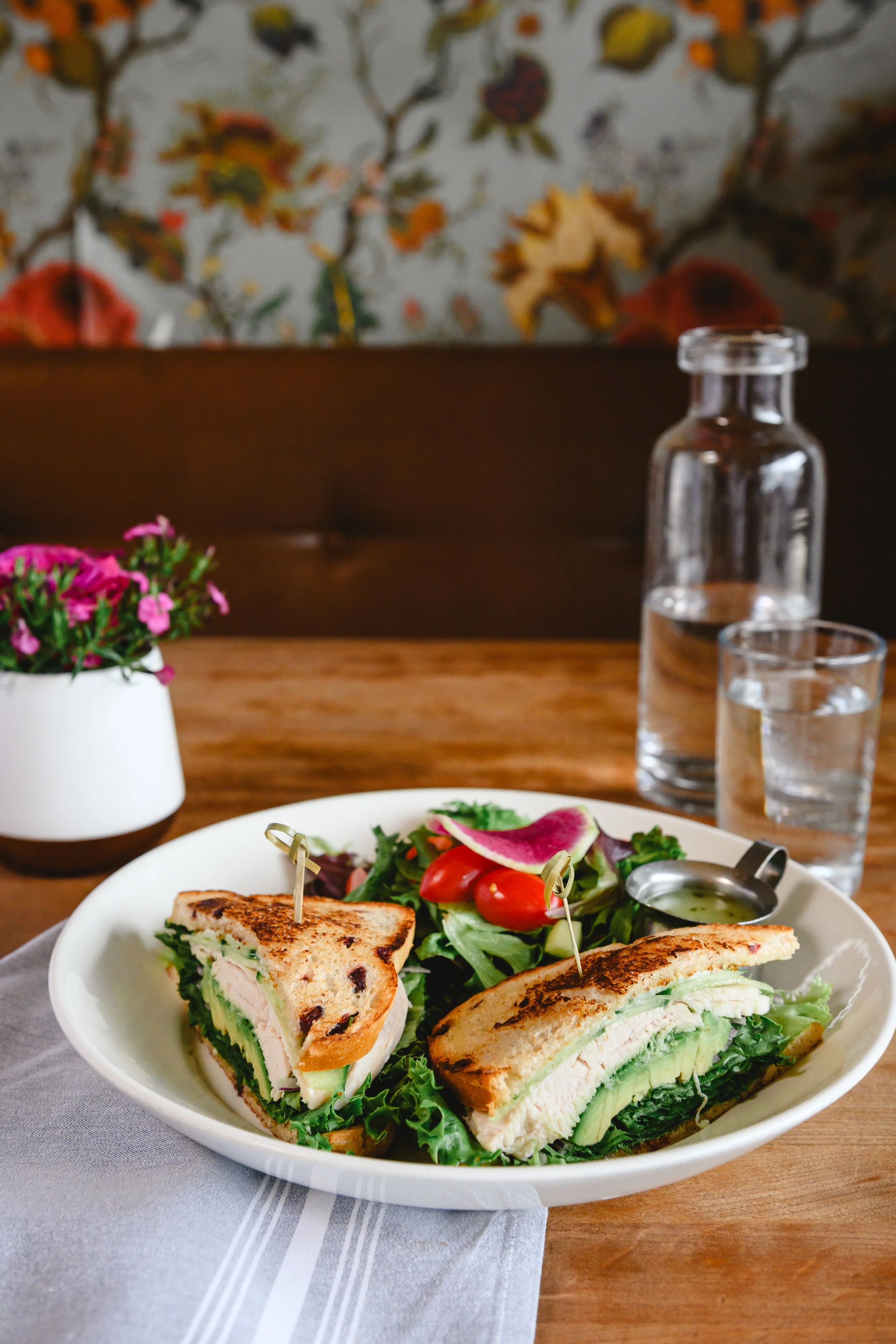 A plate with a sandwich, a side salad, a small container of dressing, a water pitcher, and two glasses of water on a wooden table, with a de