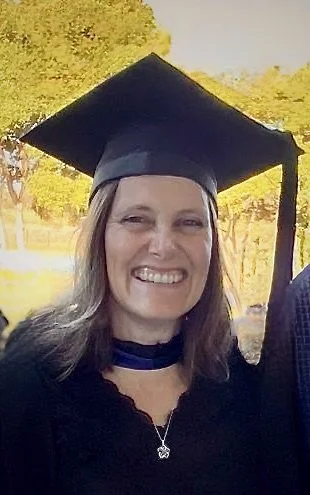 Woman wearing graduation cap smiling outdoors with trees in the background.