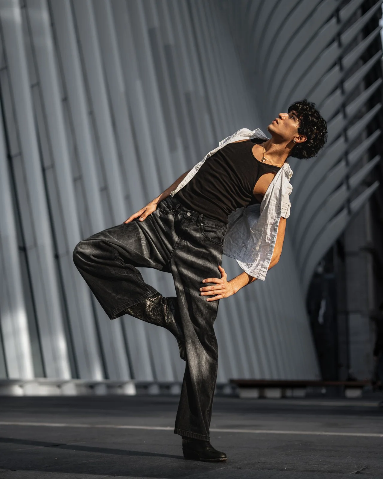 A person with short curly hair posing confidently in an urban setting with modern, curved architecture in the background, wearing black pants, a black top, and an open white shirt.