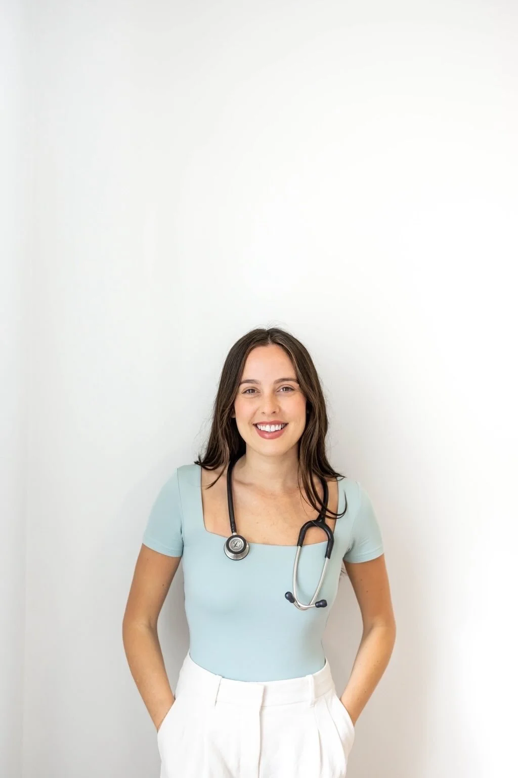 A young female healthcare professional with long brown hair, smiling, wearing a light blue top and white pants, with a stethoscope around her neck, standing against a plain white background.