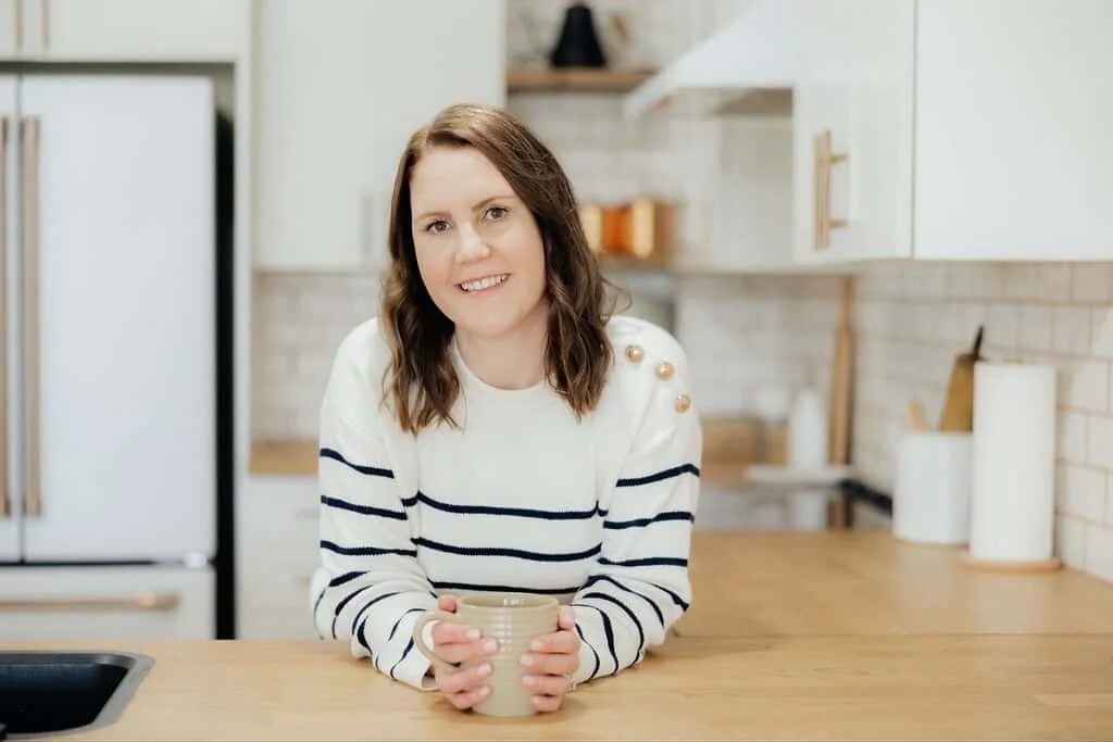 woman with shoulder-length brown hair smiling while holding a mug in a modern kitchen