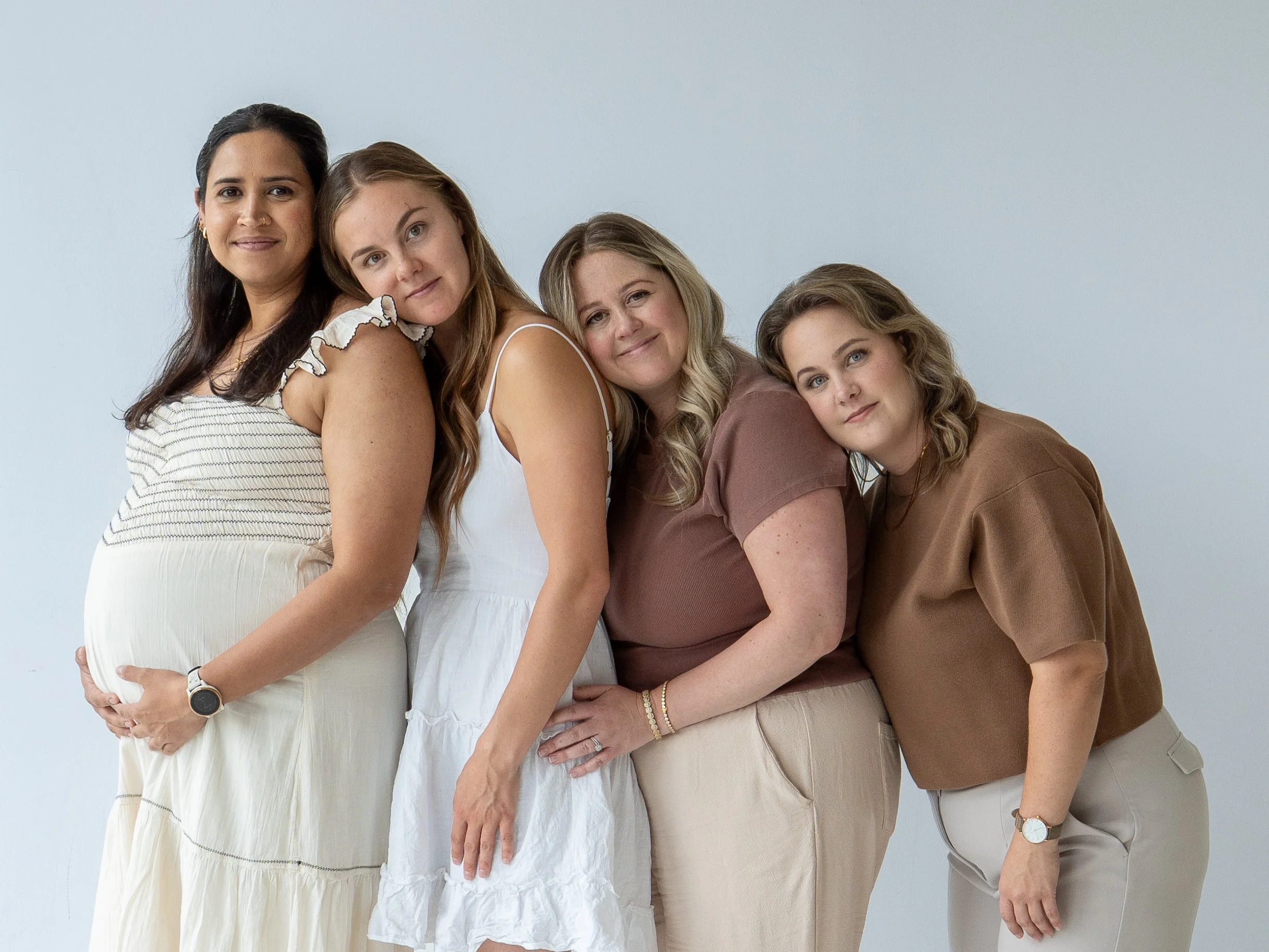 Four women of different ethnicities, including pregnant woman, standing closely together, leaning on each other against a plain light-colored background, smiling gently.